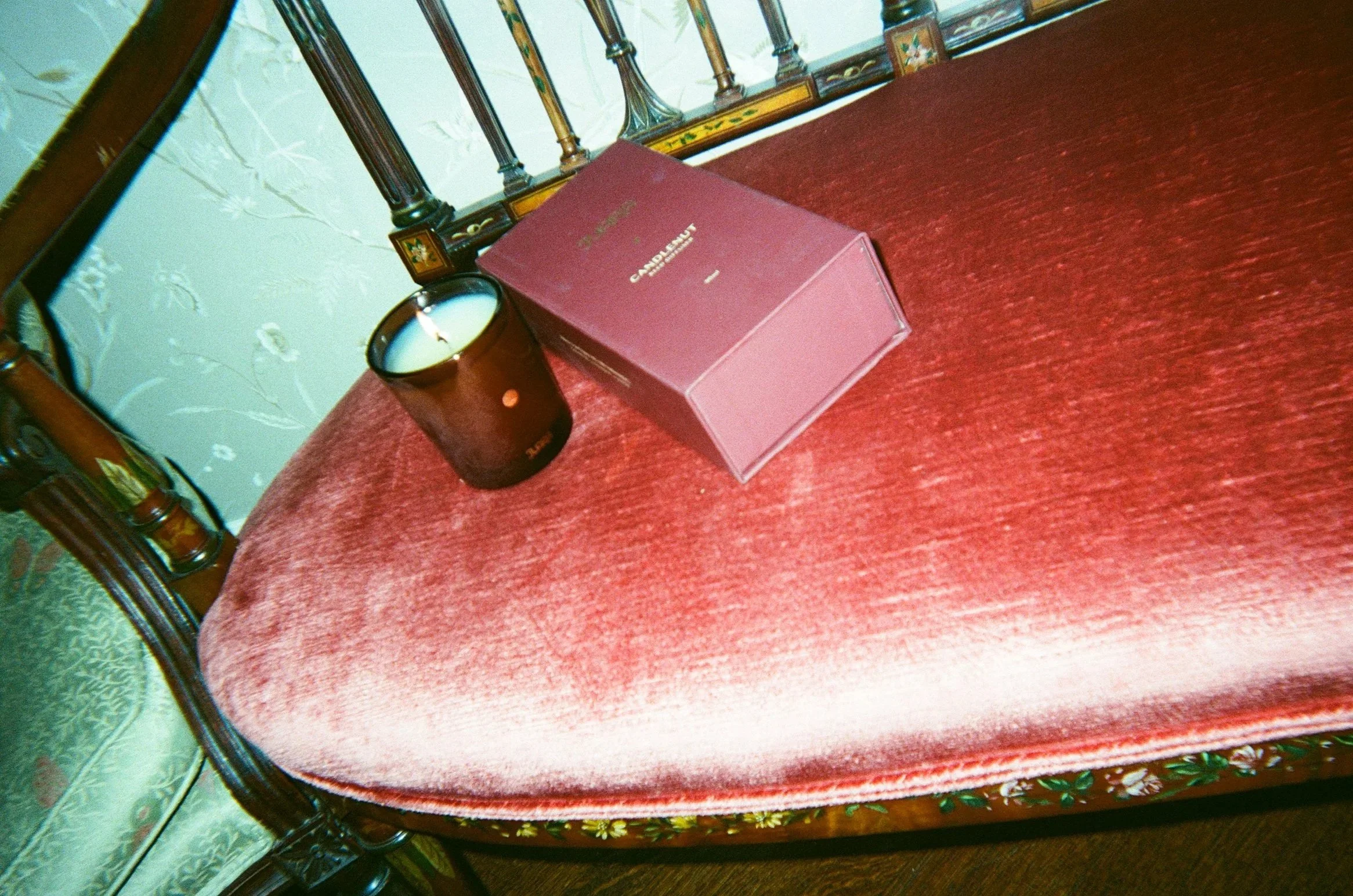 A vintage wooden chair with a pink velvet cushion, a candle in a brown glass holder, and a purple box on the cushion.
