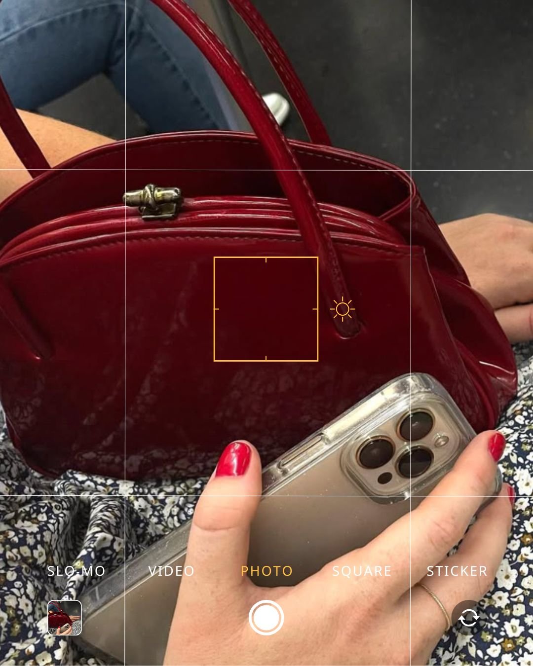 Close-up of a person holding a smartphone with a camera app open, focused on a red handbag placed on their lap. The person's nails are painted red, and they wear a gold ring. The handbag has handles and a zipper, and the background shows a person sit