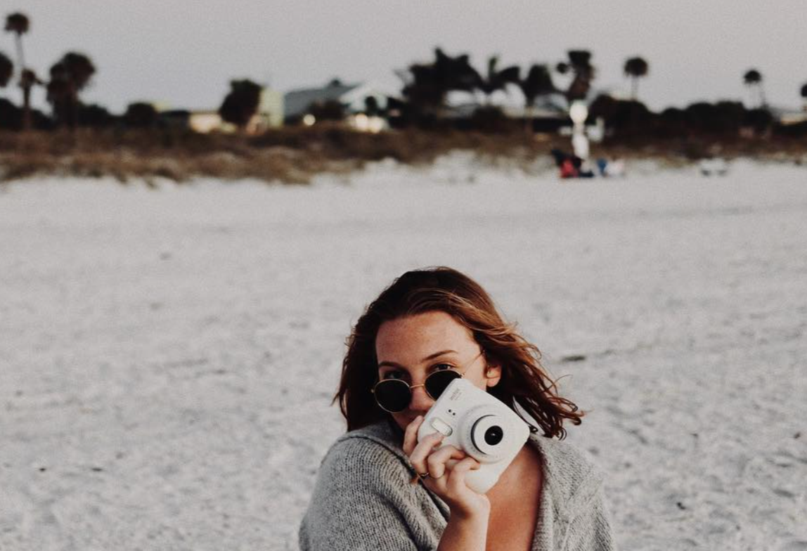 Woman with short brown hair wearing sunglasses and a gray sweater, holding a Polaroid camera, at a beach with sand and palm trees in the background.