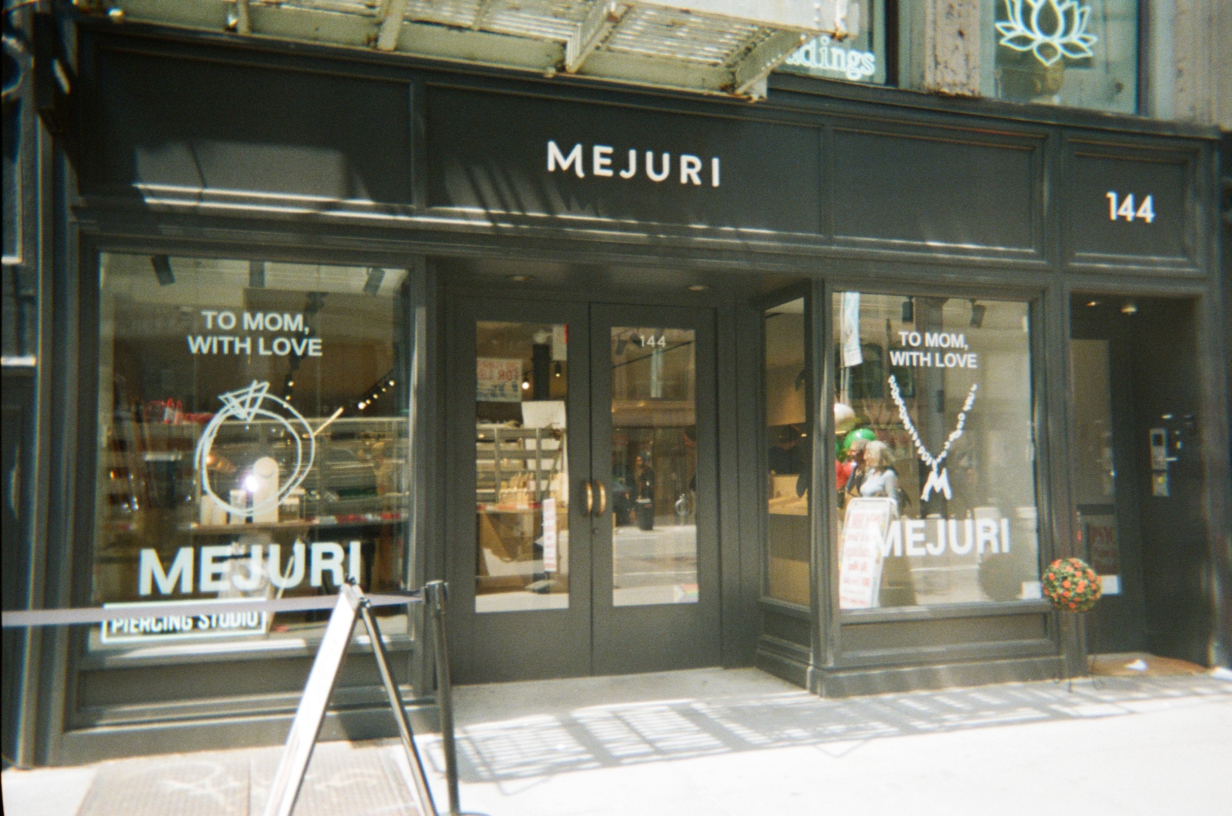 The storefront of a jewelry store named MEJURI with large glass windows and black framing. Two white signs on the windows read "TO MOM, WITH LOVE." The store's name is displayed above the door and on the windows. The store number 144 is visible on th