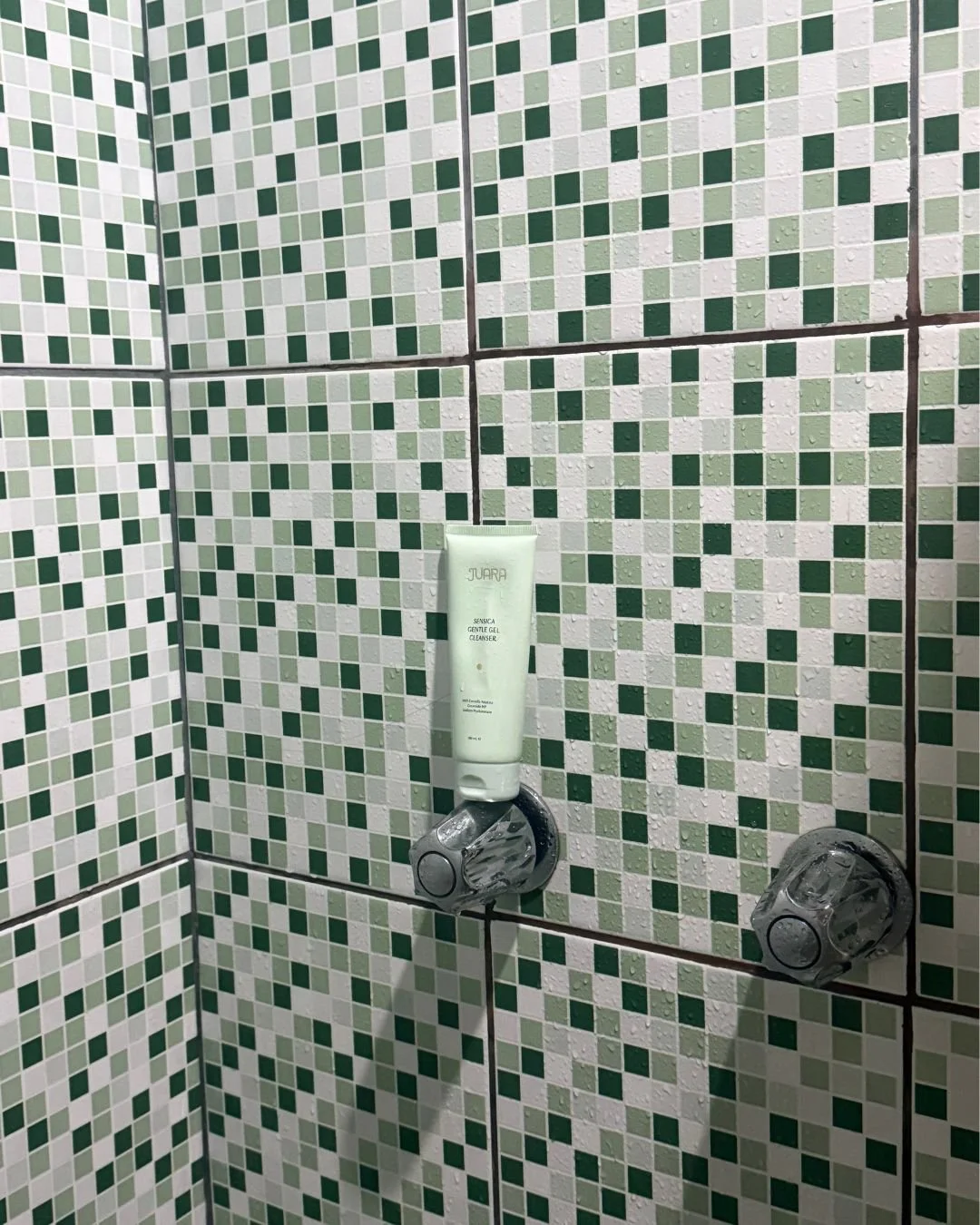 A close-up of a wall with green and white mosaic tiles in a shower, featuring a white gel cleanser tube mounted on a soap holder, with two clear plastic cups attached to the wall.