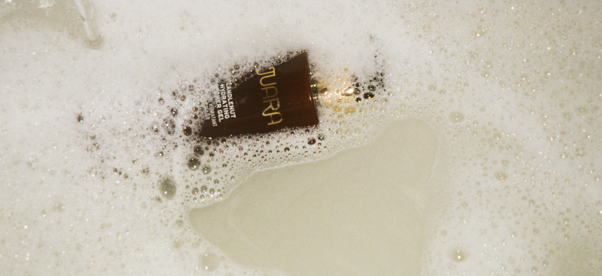 A dark brown tube of TURA skincare product partially submerged in soapy water with white foam bubbles.