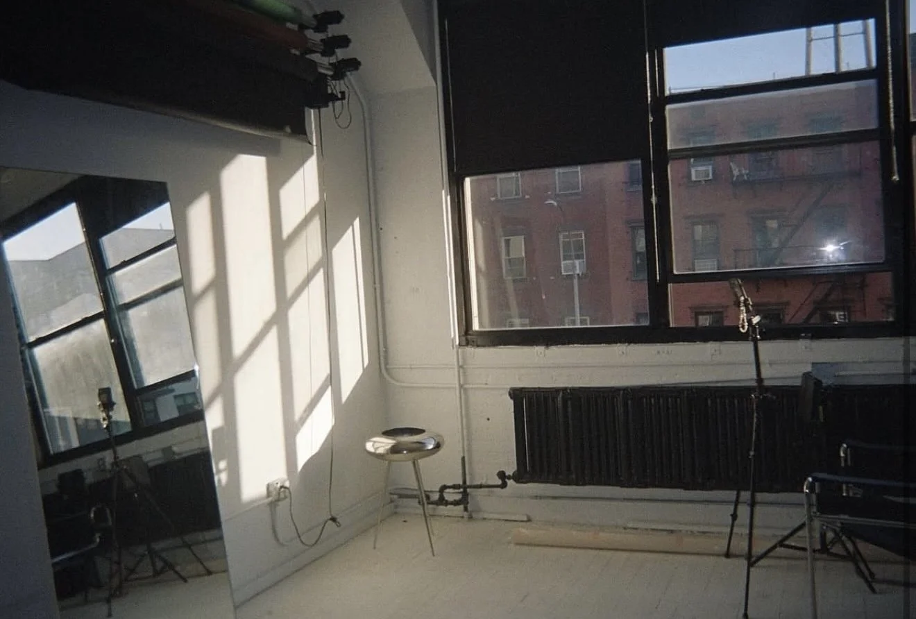 An empty room with large open windows casting shadows onto the beige floor. A stool, a mirror, and some equipment are visible.