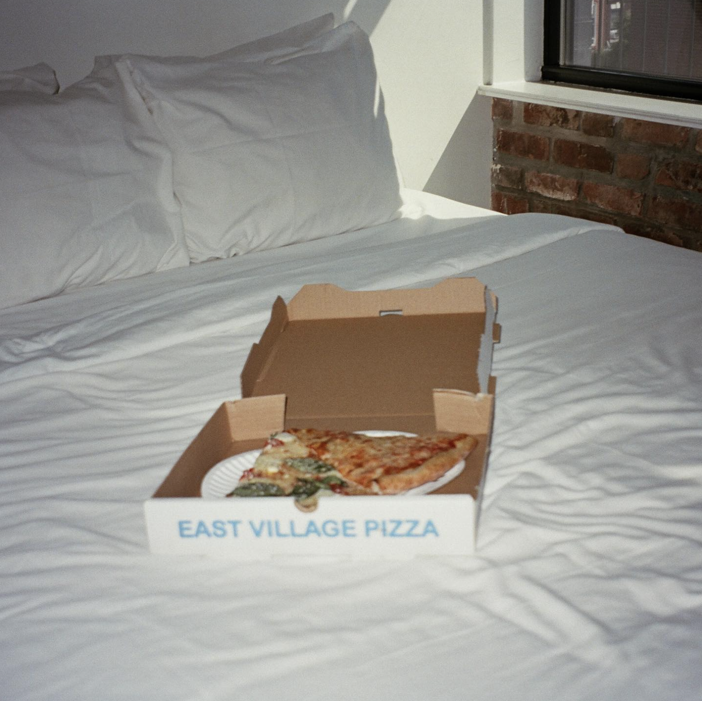 Open pizza box with two slices of pizza on a white paper plate on a bed with white sheets and pillows, near a window with brick wall detail.