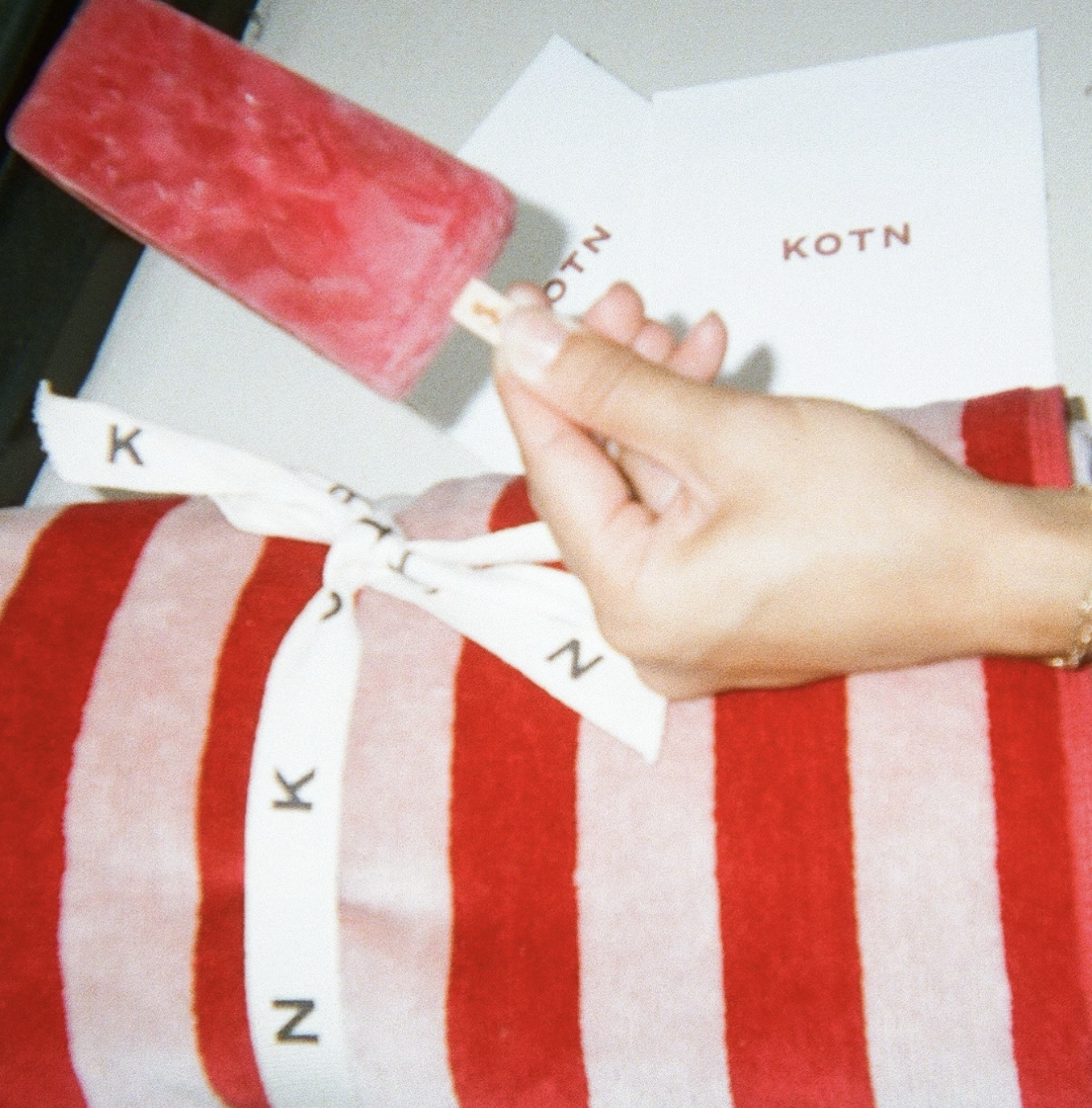 Person holding a pink popsicle in front of a white box with 'KOTN' written on it, wearing a red and pink striped sweater.