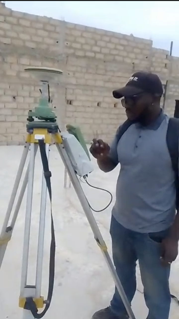 Un homme portant un chapeau noir, lunettes et un sac à dos, se tenant à côté d'un niveau topographique sur un trépied, à l'extérieur devant un mur en briques.