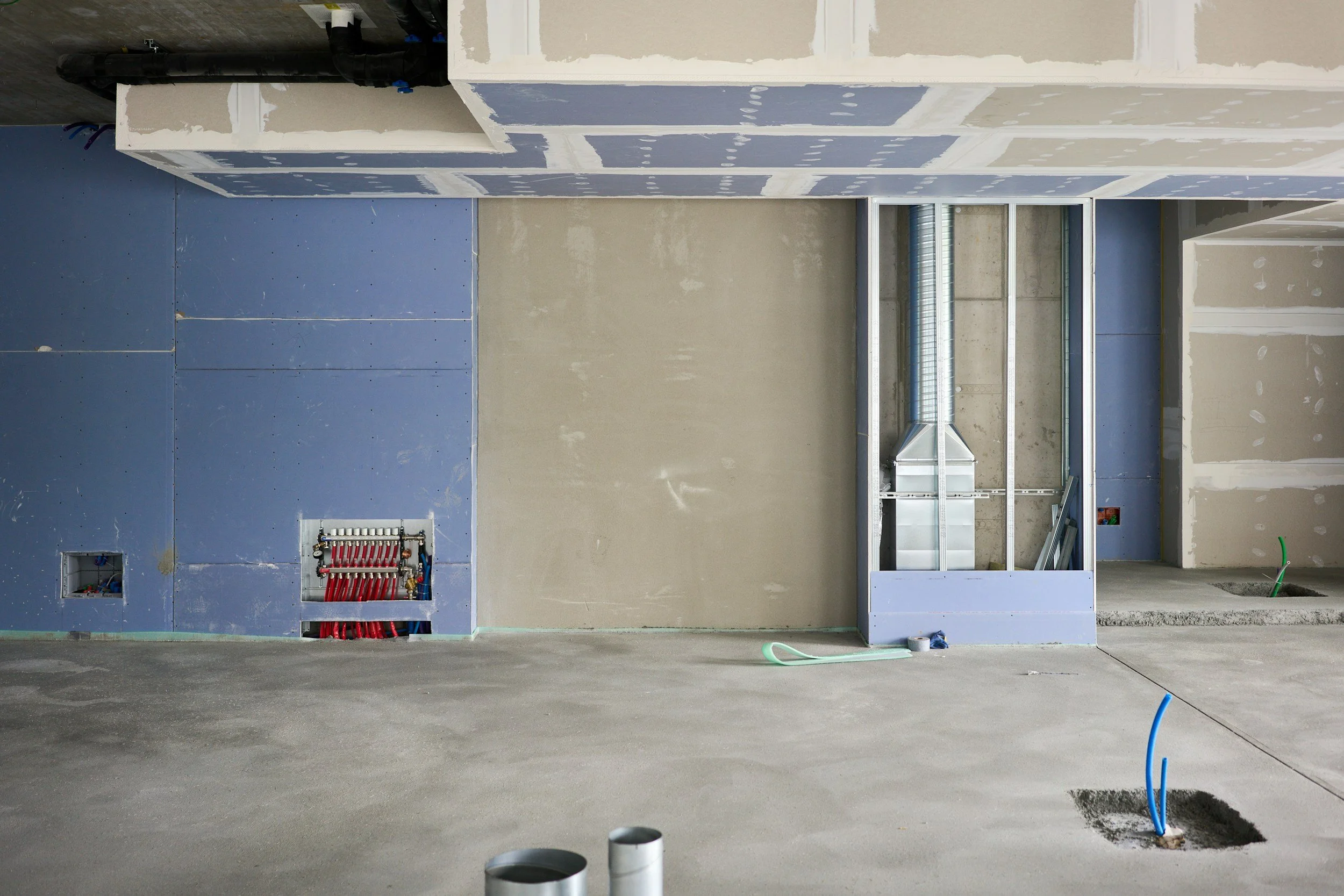 Interior of a building under construction with drywall and ductwork. Blue and beige walls, exposed pipes, and unfinished flooring are visible.