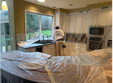A person working in a kitchen under renovation, with plastic covering the counters and bed, and a sliding glass door leading outside.