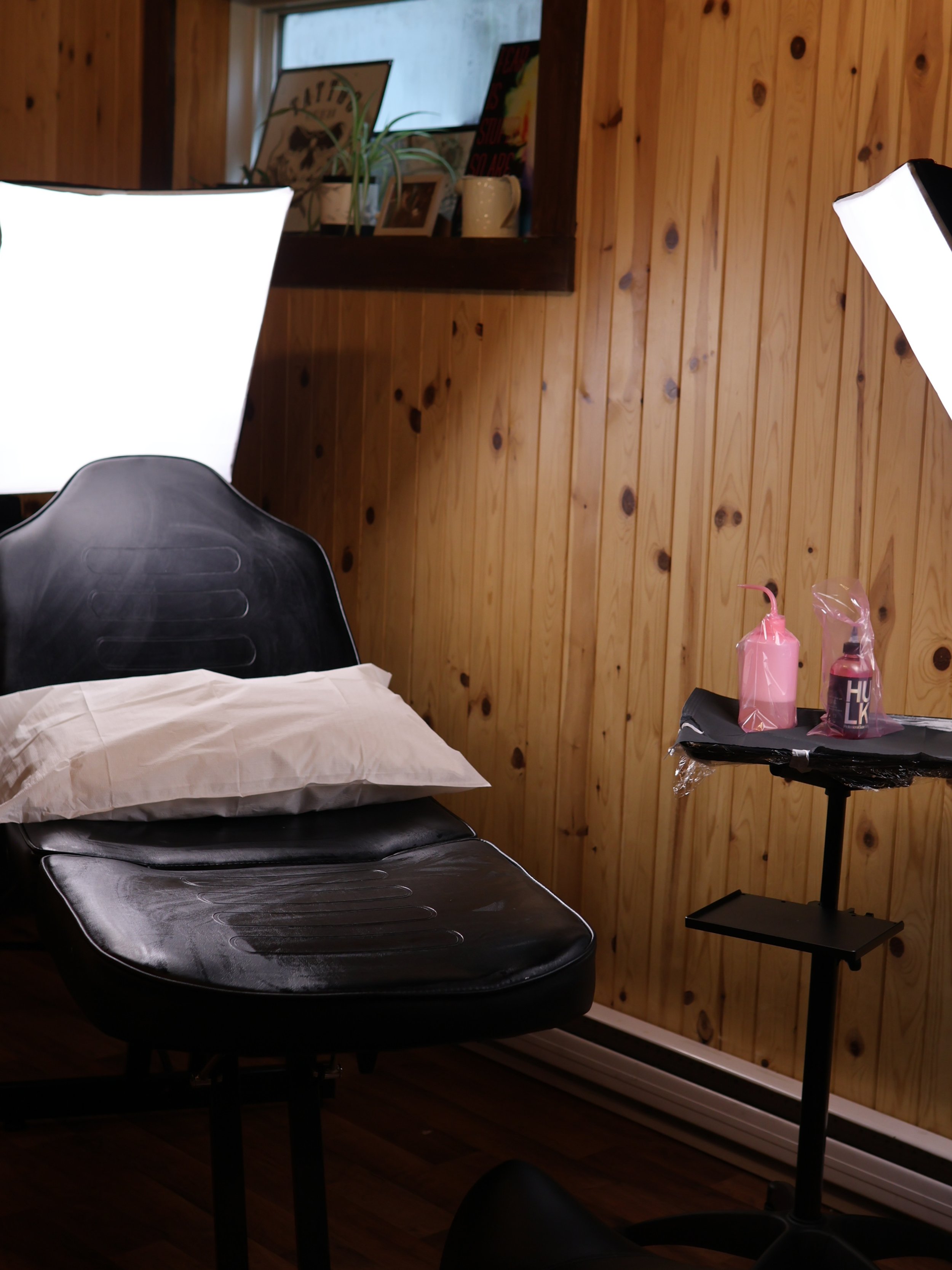 Studio de tatouage avec un lit d'examen en cuir noir, un coussin, et une table avec deux bouteilles de produits pour tatouage. Éclairage de studio professionnel avec plusieurs lumières.