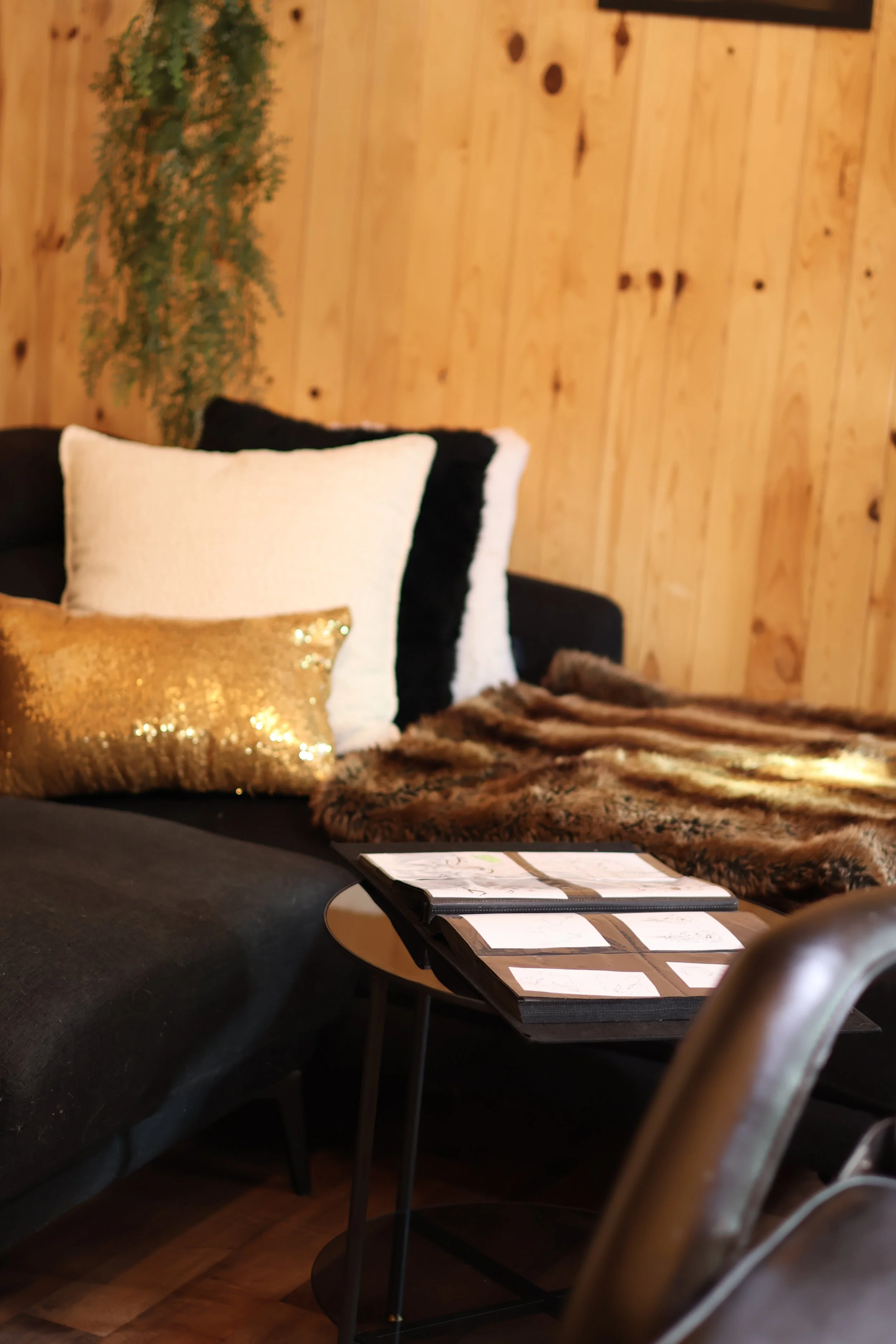 Un canapé noir avec plusieurs coussins, dont un doré à sequins, un blanc, et un noir, avec une couverture en fourrure, devant un mur en bois. Un petit meuble noir avec des albums ou livres ouverts est placé devant le canapé.