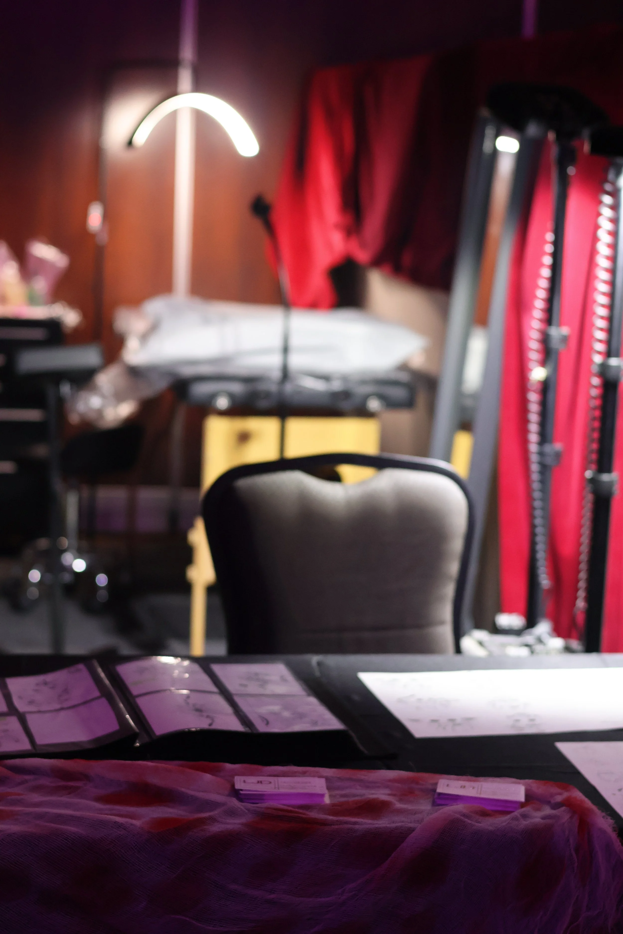 Une salle de théâtre ou studio de répétition avec une chaise au centre, des documents sur une table, un lampadaire, et des éléments d'éclairage ou de décor en arrière-plan.