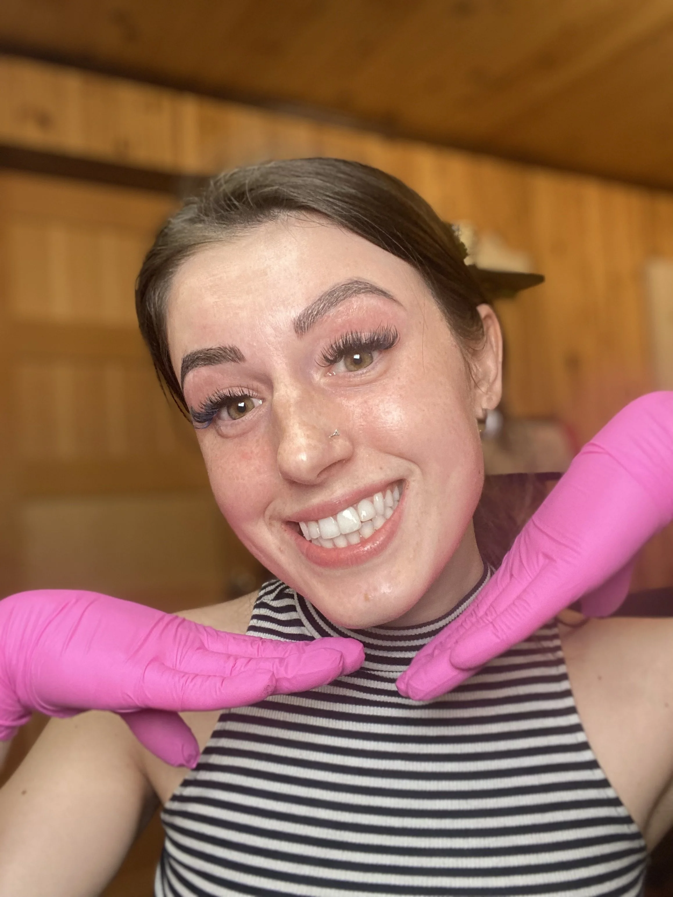 Jeune femme souriante avec un maquillage prononcé, portant des gants roses et un haut à rayures noires et blanches, dans un intérieur en bois.