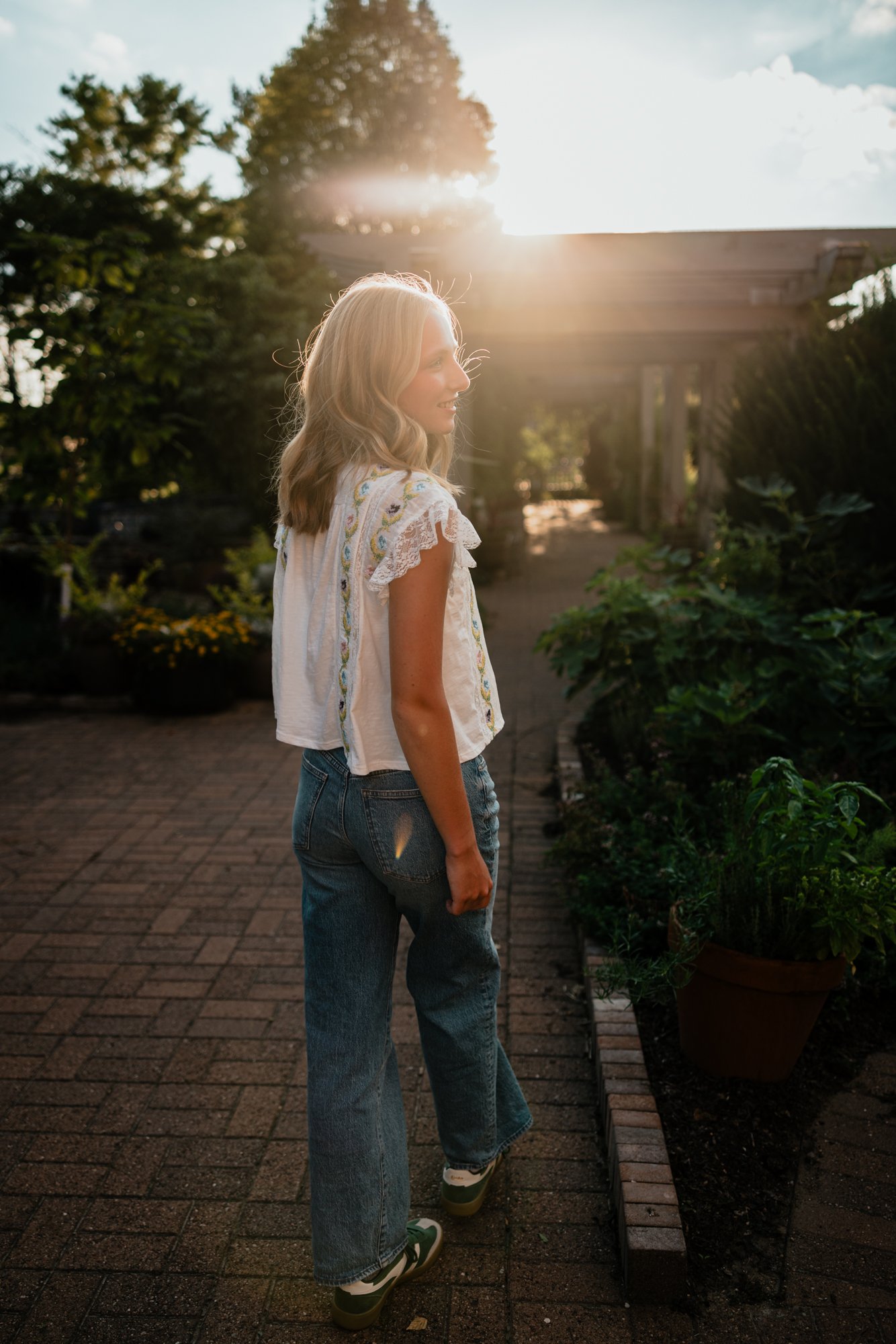 A woman with blonde hair in wavy style standing on a brick pathway in a garden during sunset, wearing a white embroidered sleeveless top, blue jeans, and sneakers, surrounded by plants and trees.