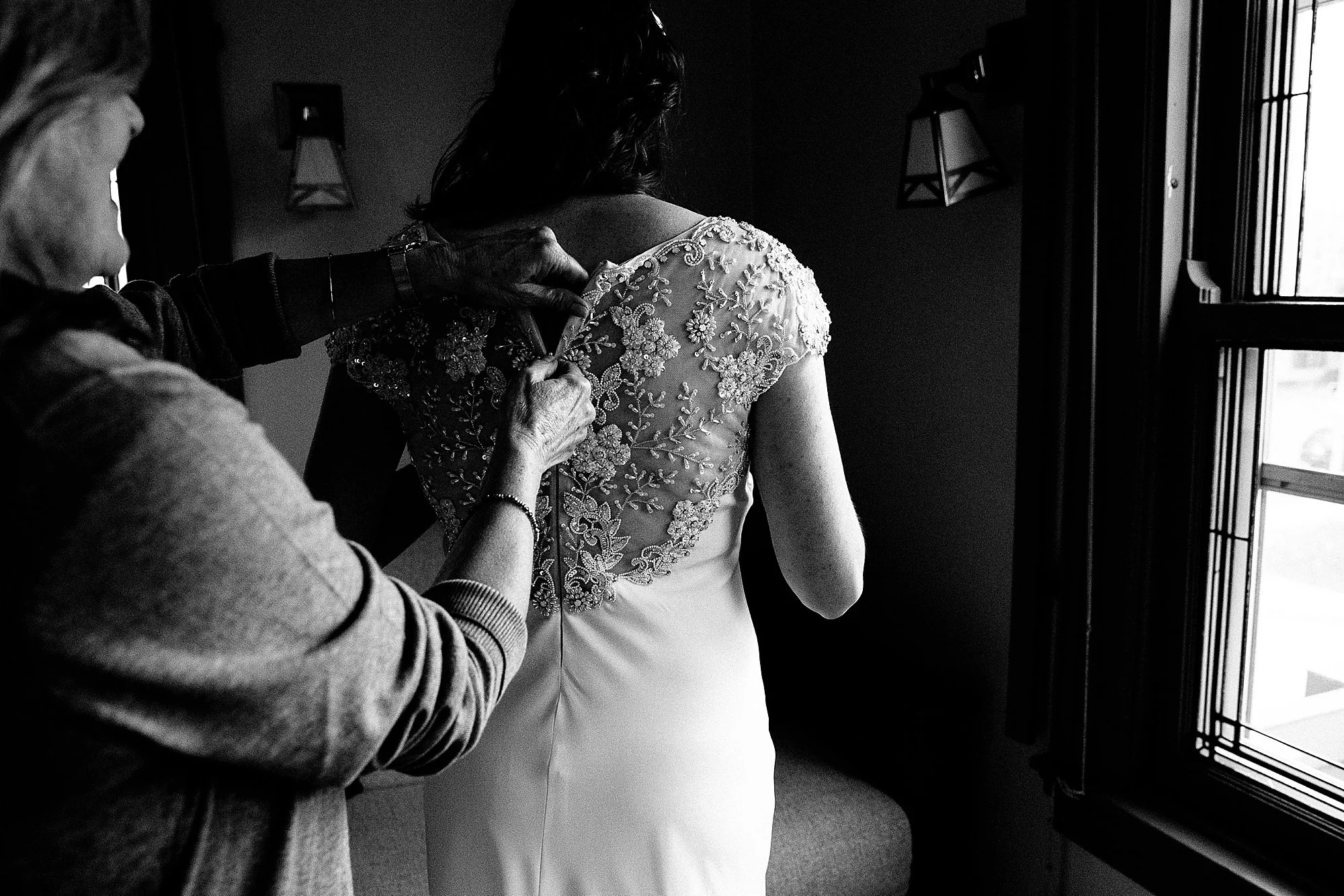 A woman helping another woman put on a wedding dress with detailed lace and floral embroidery, near a window.