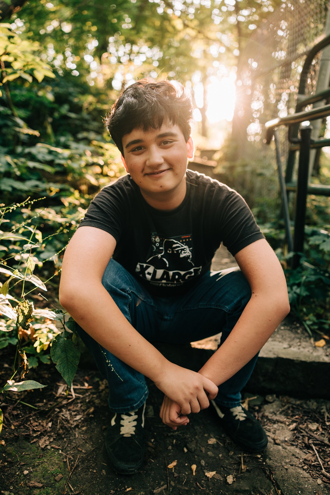 A young man crouching outdoors in a natural setting during the golden hour, with sunlight shining through trees in the background.