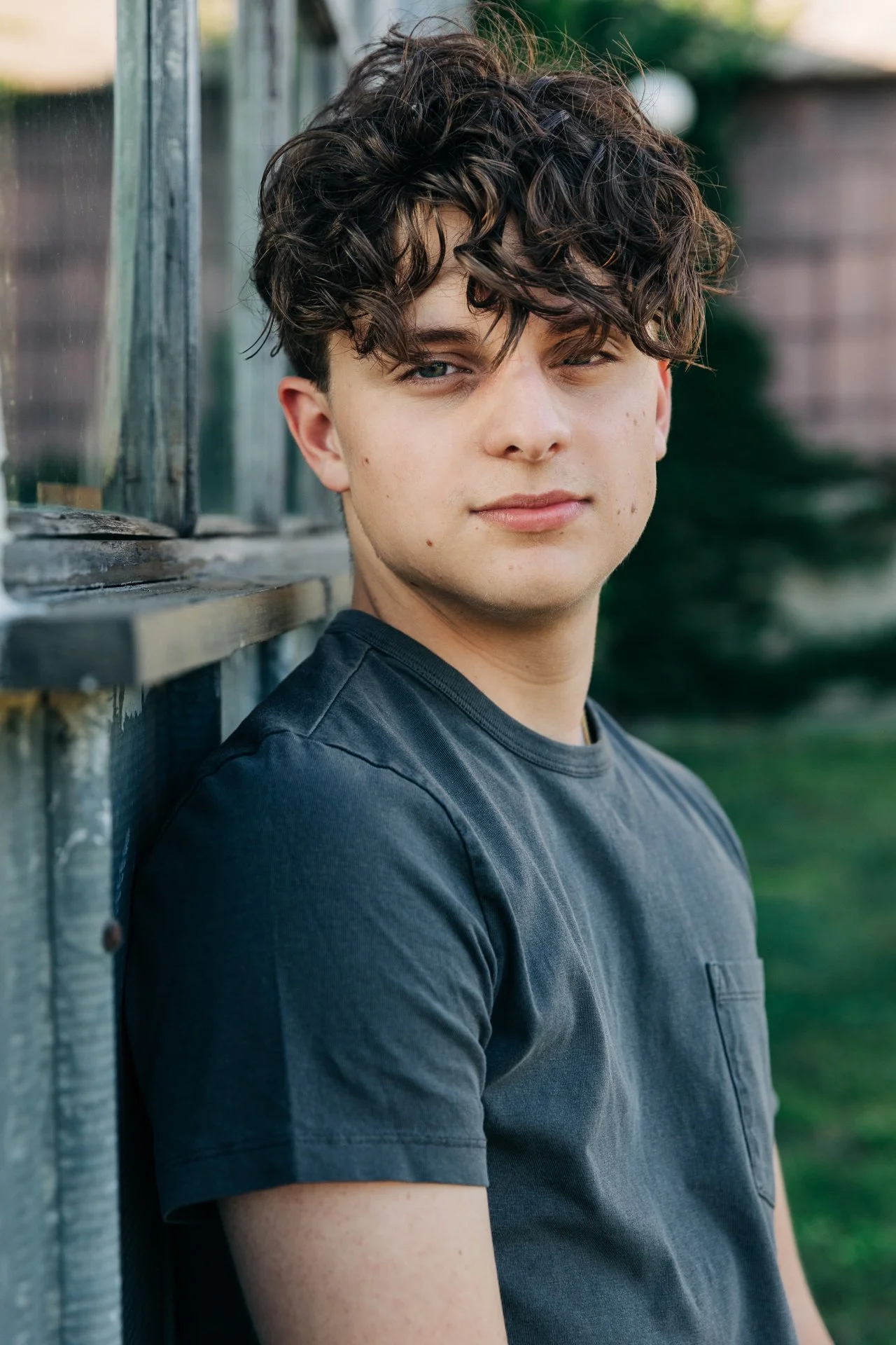 Young man with curly brown hair leaning against a wooden wall outdoors, looking at the camera with a slight smile.