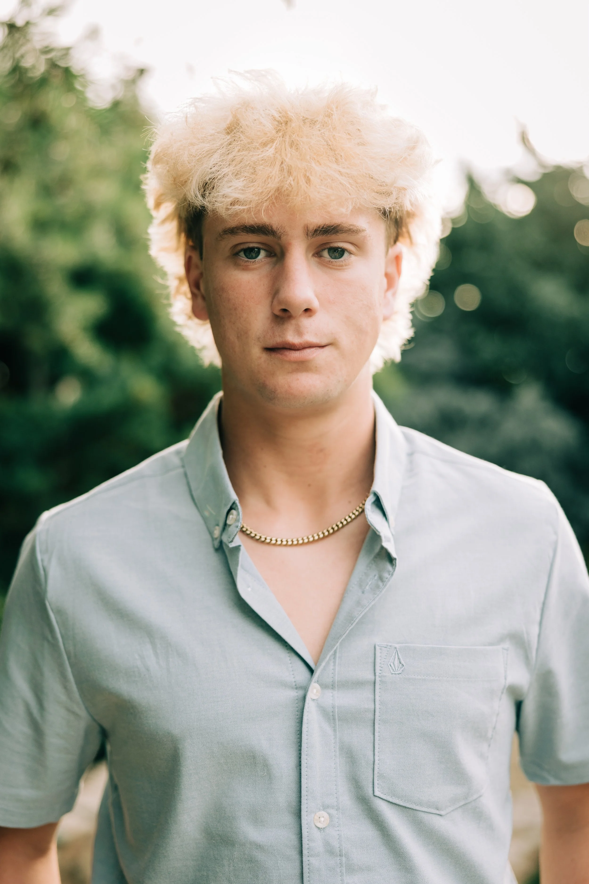 A young man with platinum blonde curly hair and blue eyes outdoors, wearing a light green button-up shirt and a gold chain necklace.