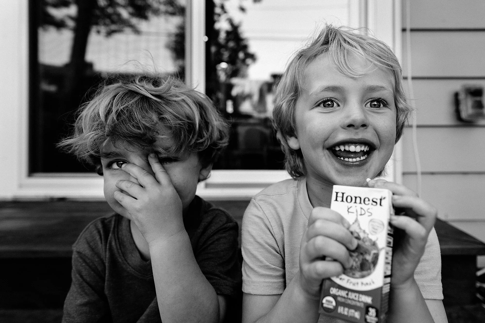 Two young boys sitting on a porch, one with curly hair covering part of his face and the other smiling broadly, holding a box of Honest Kids juice boxes.