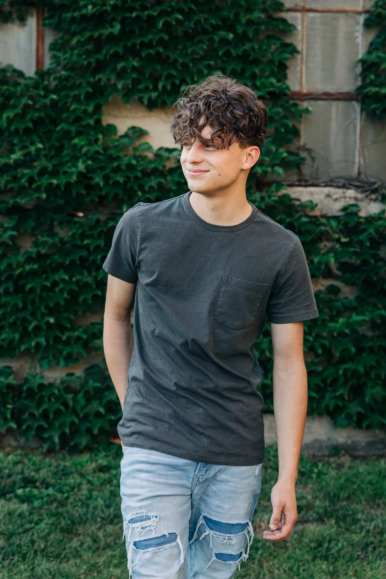 A young man with curly brown hair wearing a dark gray t-shirt and ripped light blue jeans standing outdoors near a wall covered with green ivy.