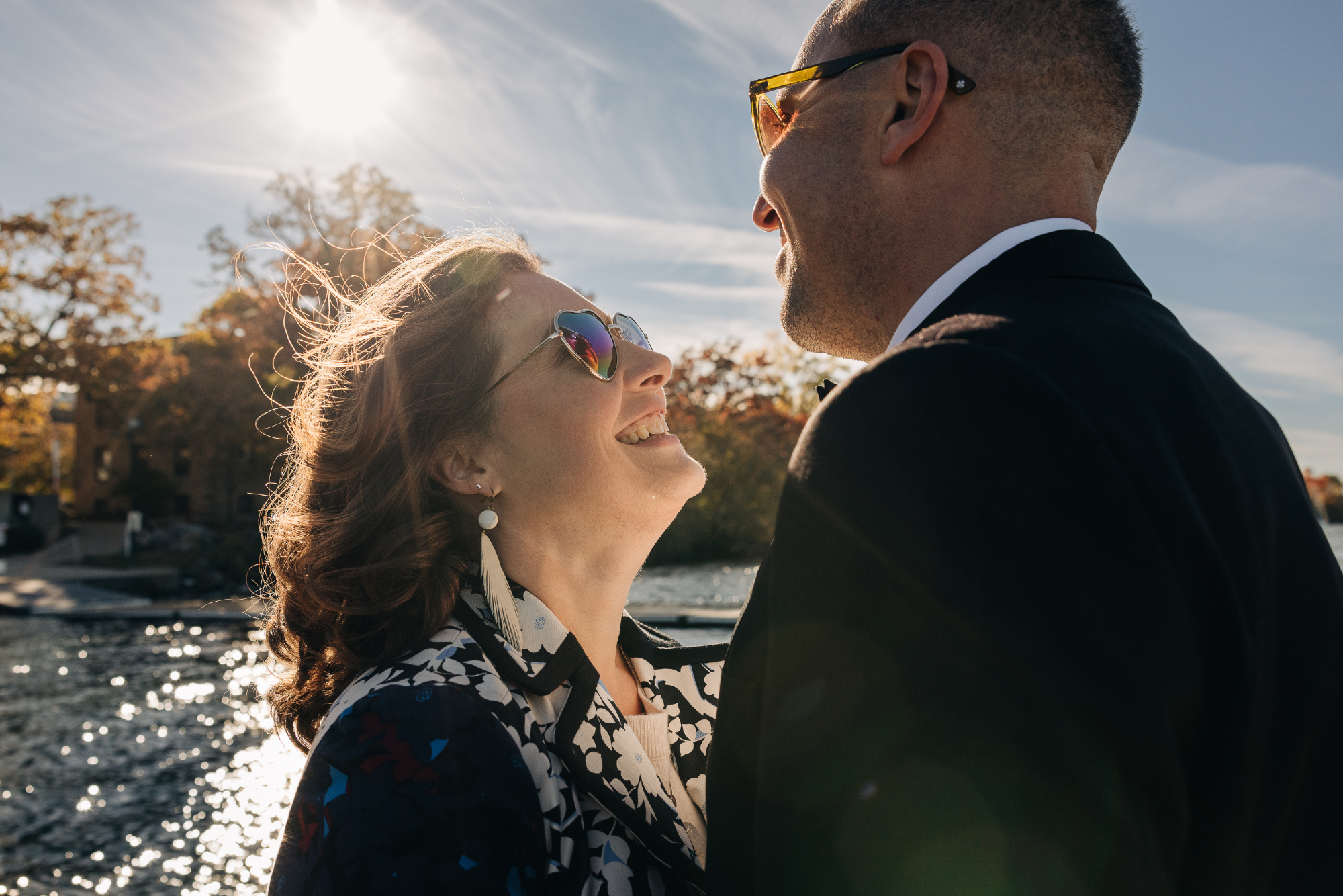 A couple smiling and looking at each other outdoors near a body of water, with trees and buildings in the background, during sunset.