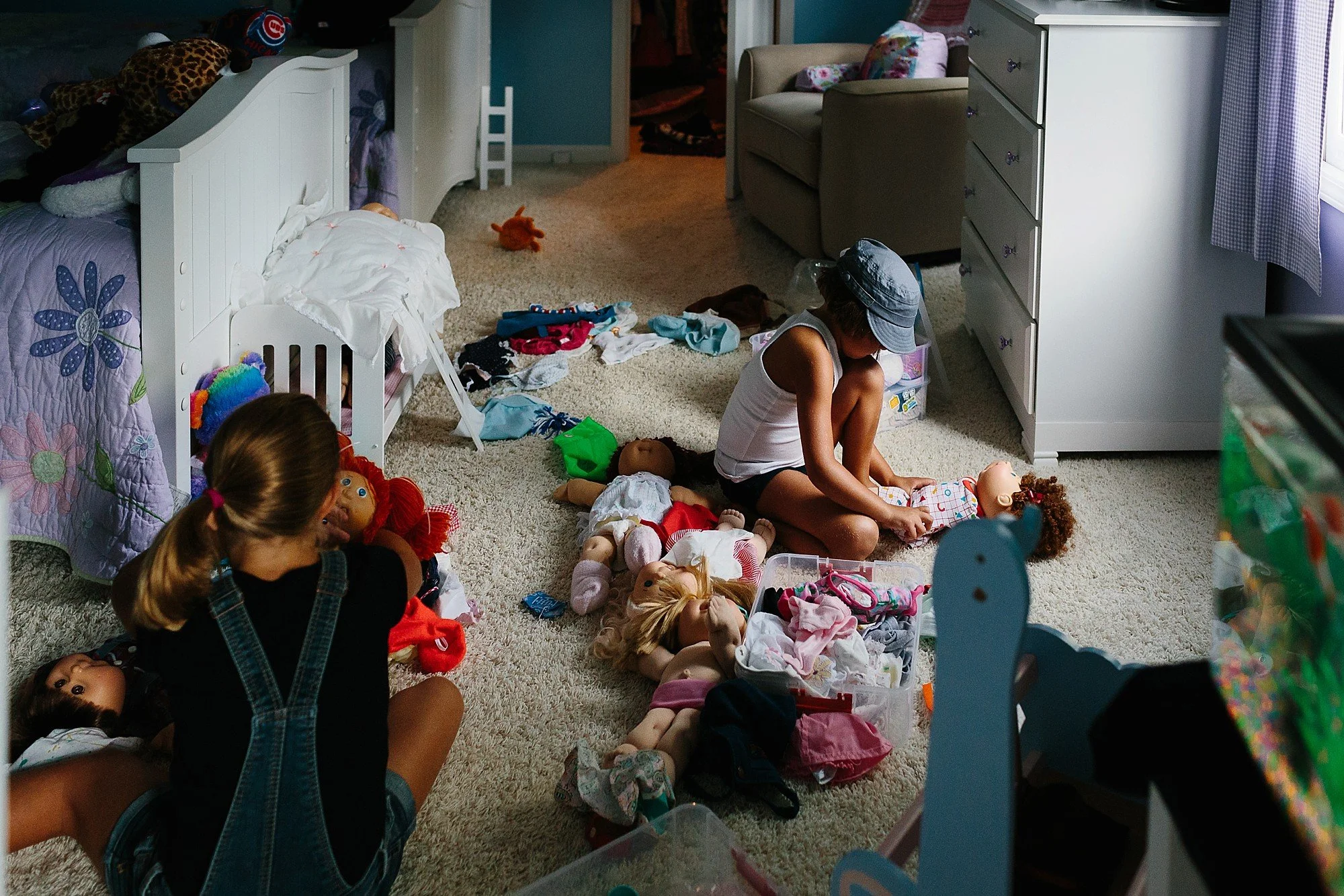 Two children playing with dolls on the carpeted floor of a cluttered bedroom, surrounded by toys, clothes, and stuffed animals.
