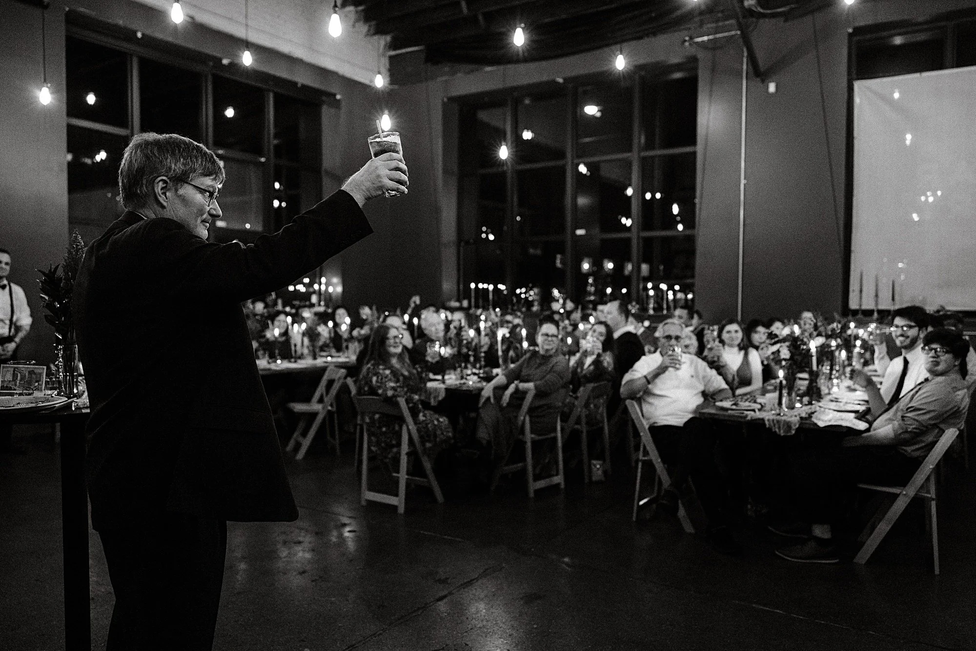 A man giving a toast at a dinner party with guests seated at tables, hanging string lights, and large windows in the background.
