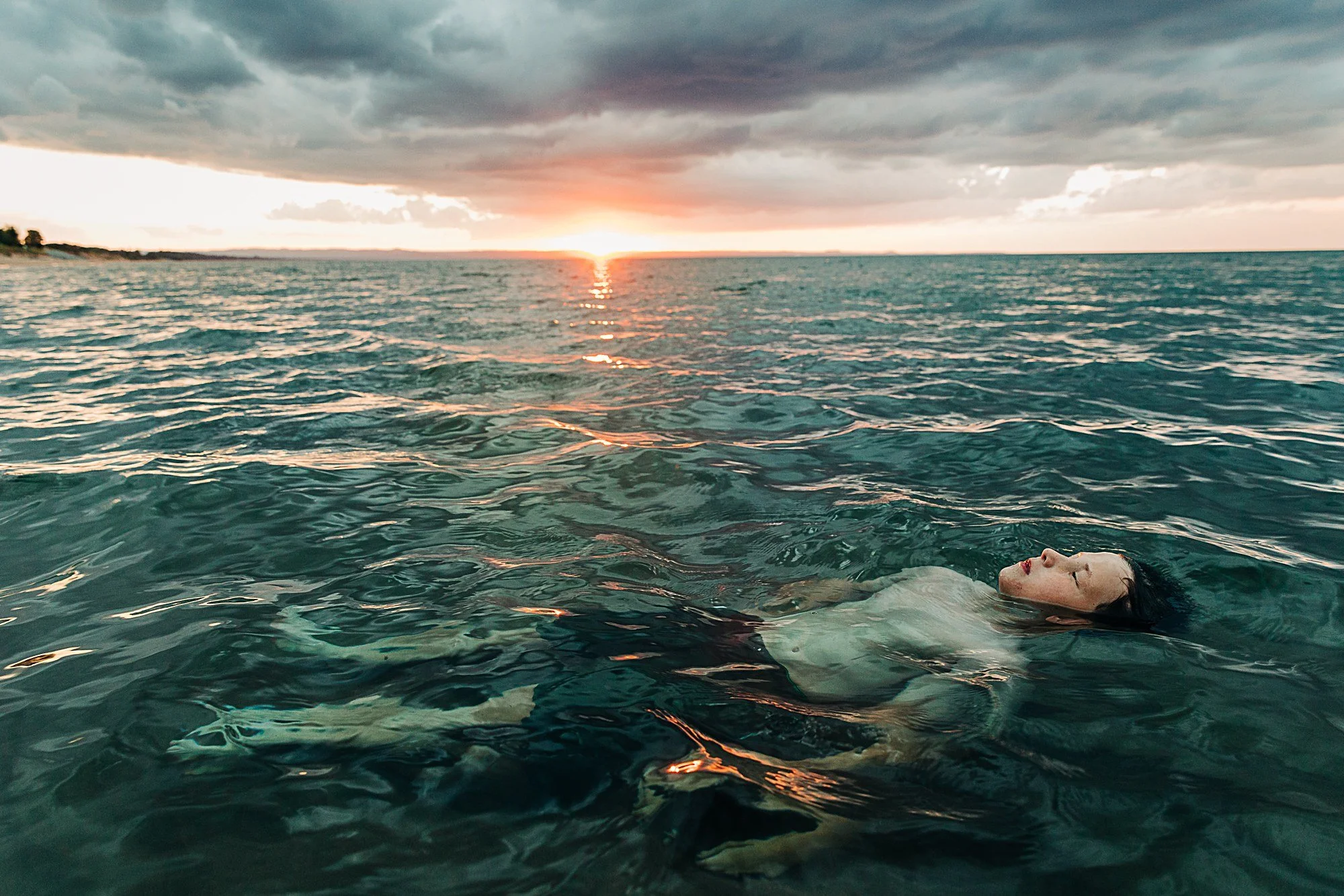 A woman floating on her back in a body of water during sunset with cloudy sky overhead.