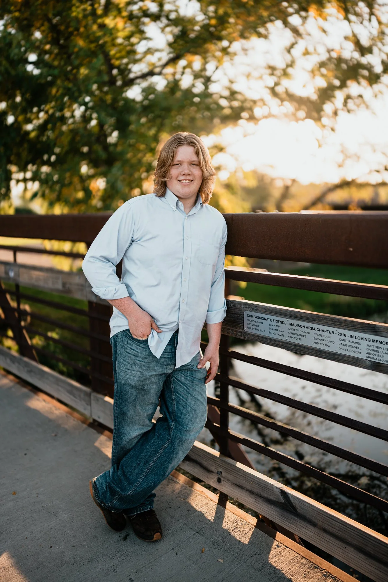 A young person with shoulder-length hair, wearing a light blue shirt and jeans, stands leaning against a wooden railing on a bridge near a river during sunset, with trees in the background.