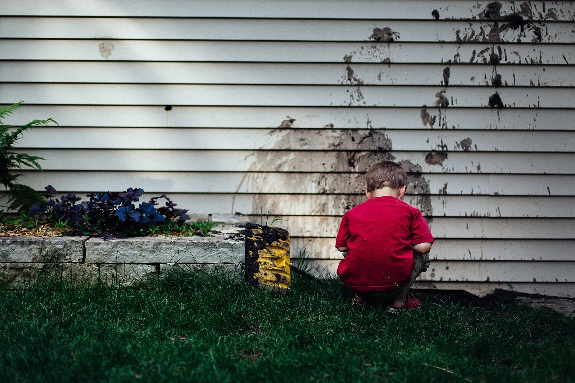 A young boy in a red shirt squats next to a dirty, soot-stained wall or siding.
