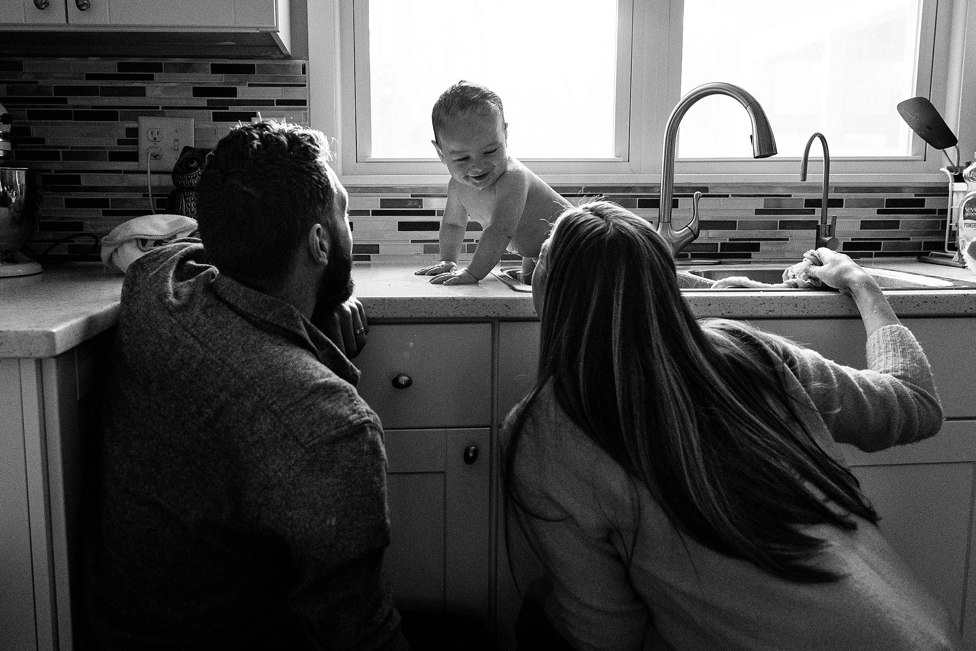 A toddler at a kitchen sink with two adults nearby, with the toddler smiling at the adults.