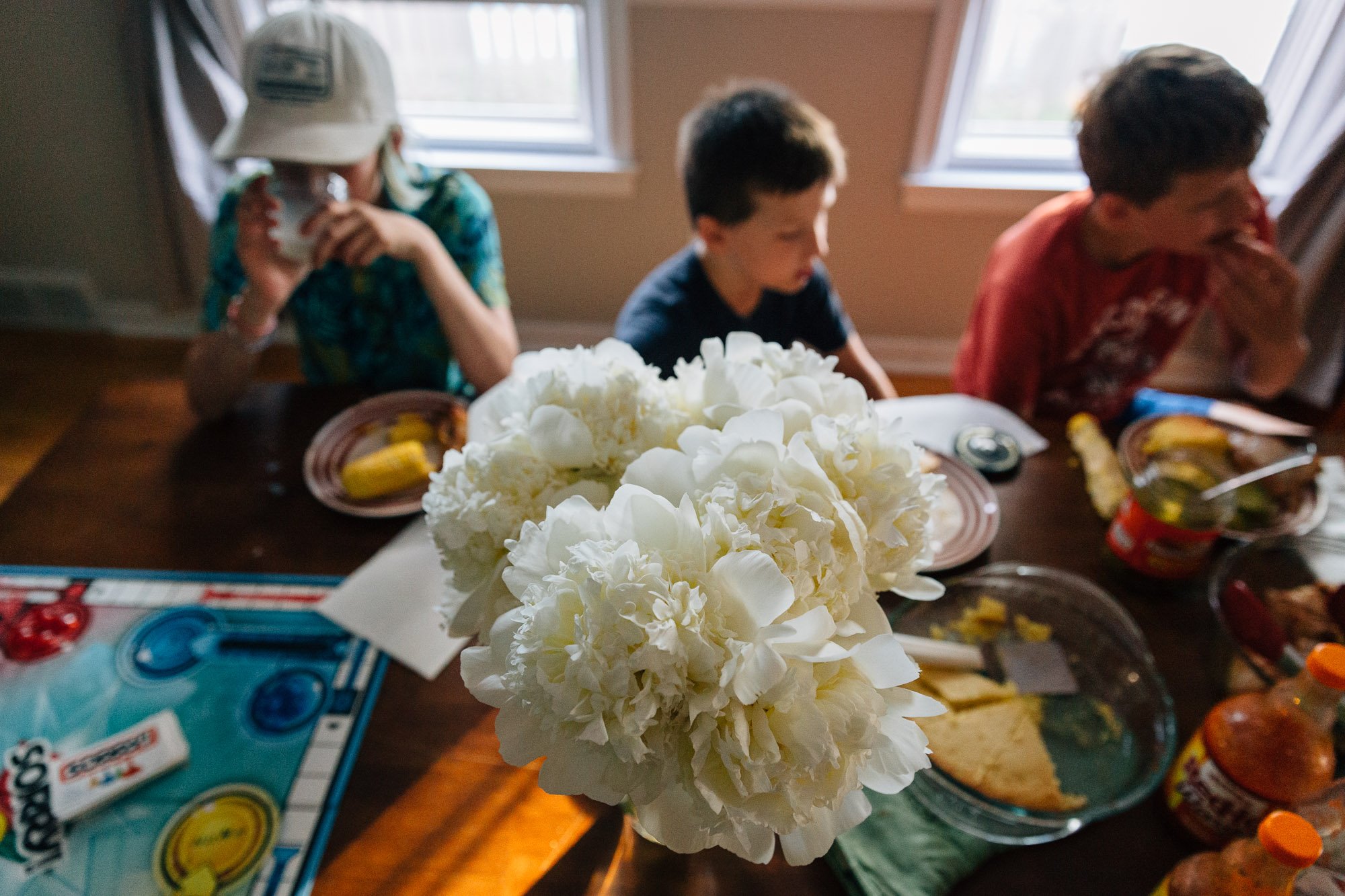 A white peony centerpiece on a table with children sitting in the background during a meal, with food, drinks, and toys visible on the table inside a room with windows.