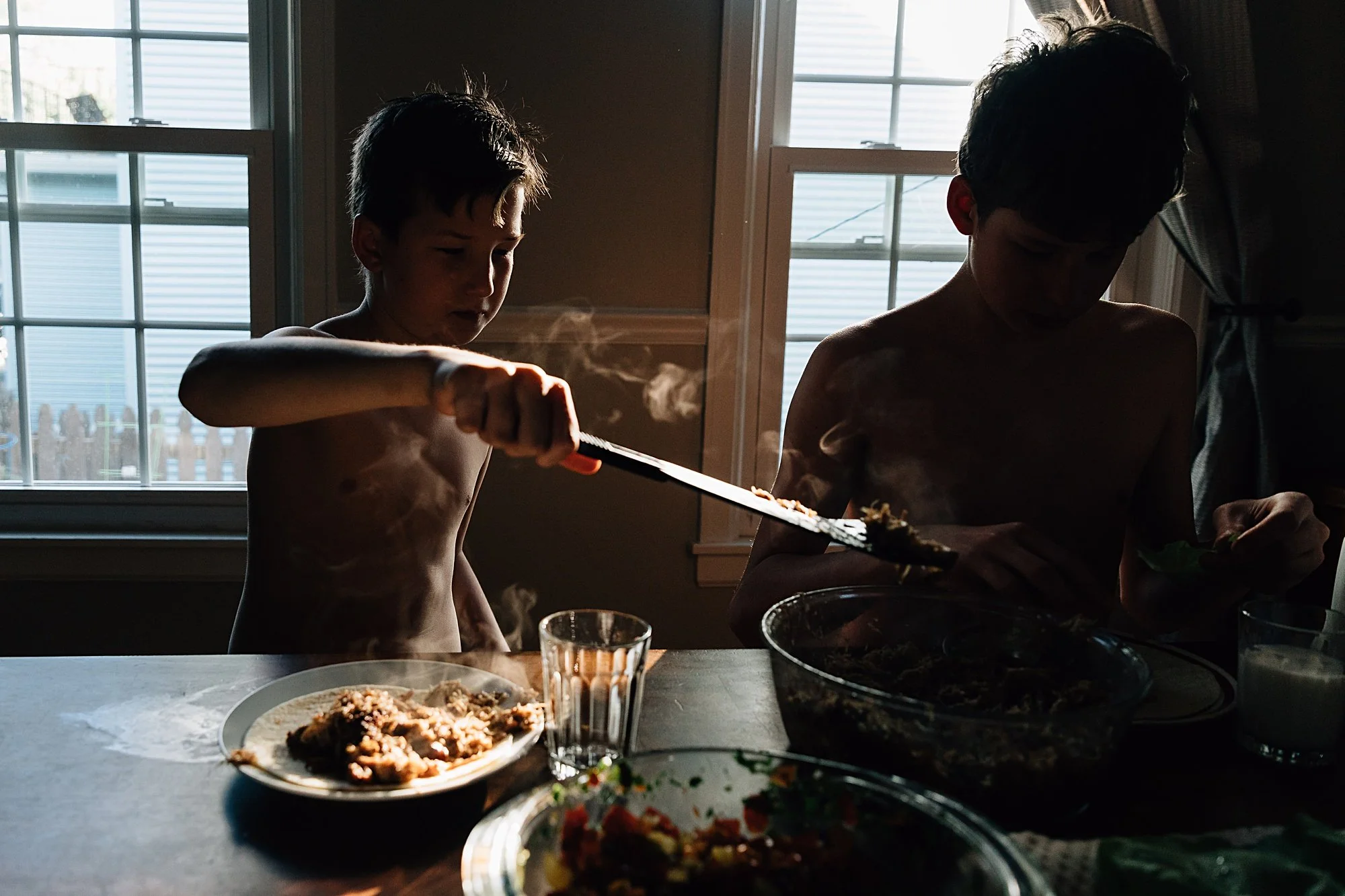 Two young boys, shirtless, preparing food at a table indoors with natural light coming through the windows.