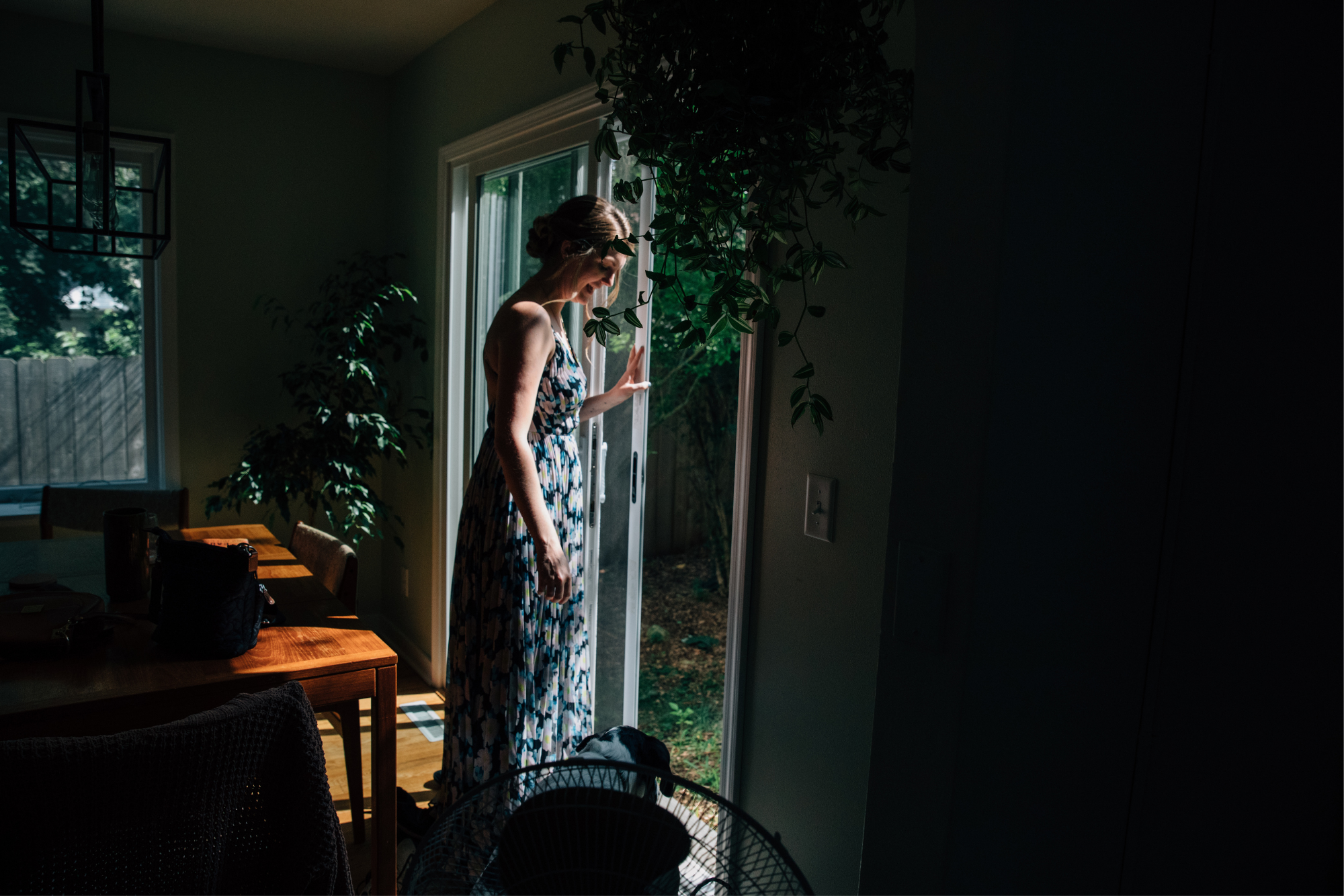 A woman in a patterned dress standing by a sliding glass door, looking outside into a garden with sunlight illuminating her face and green foliage.