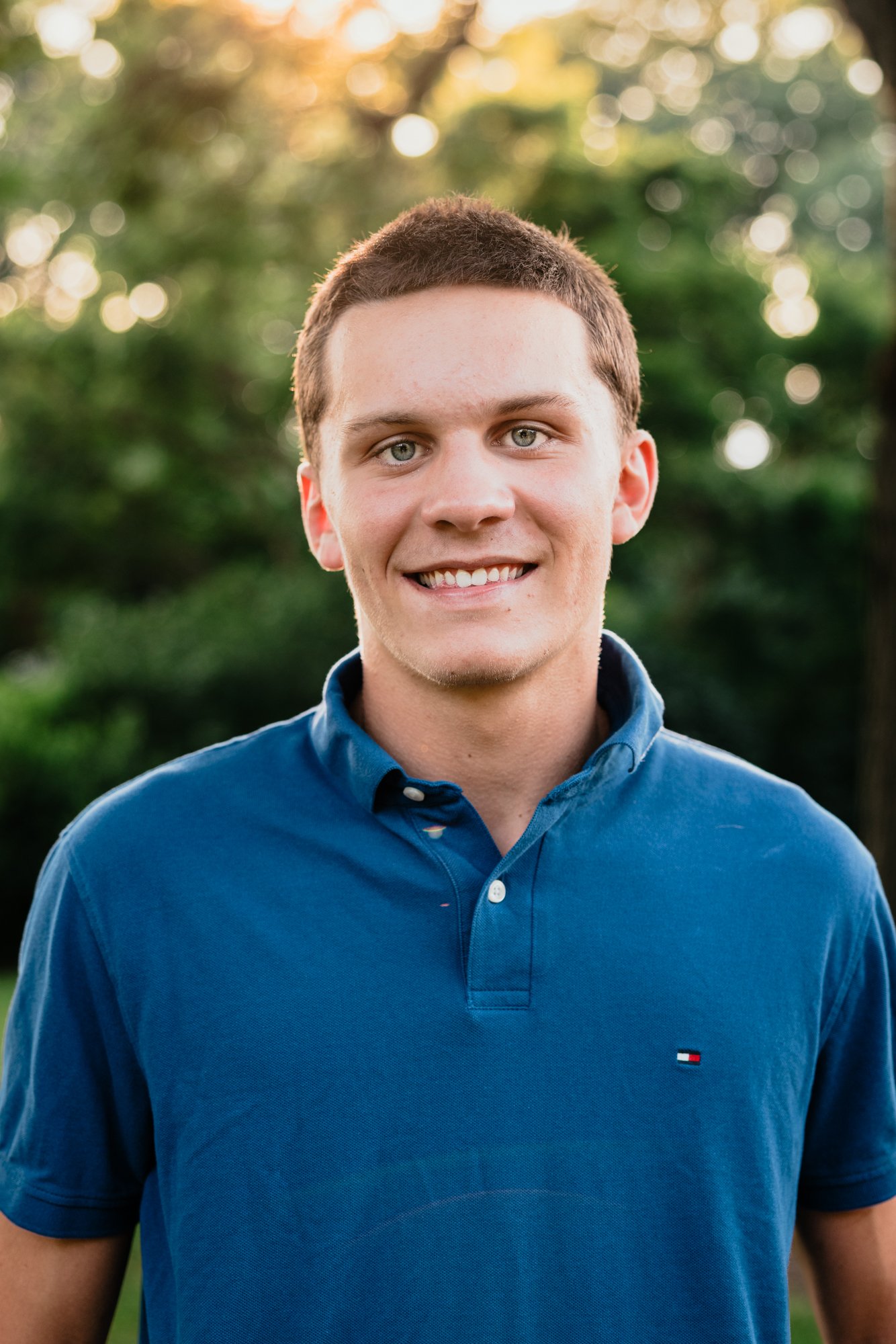 A young man with short brown hair and blue eyes smiling outdoors, wearing a blue polo shirt.