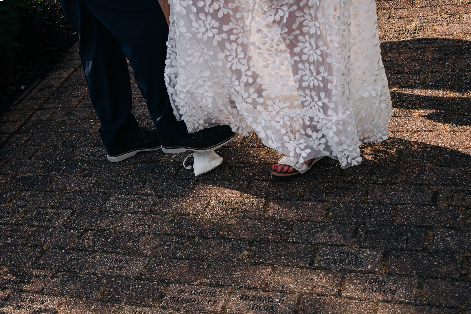 Close-up of a bride and groom standing on a brick pathway, with the bride wearing white flower-patterned dress and sandals, and the groom in dark trousers and black shoes.