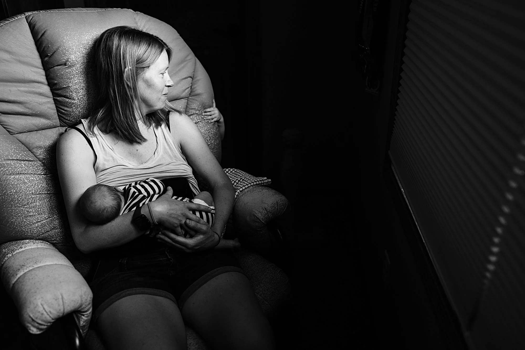 A woman sitting on a sofa, breastfeeding a baby while watching television in a dark room.