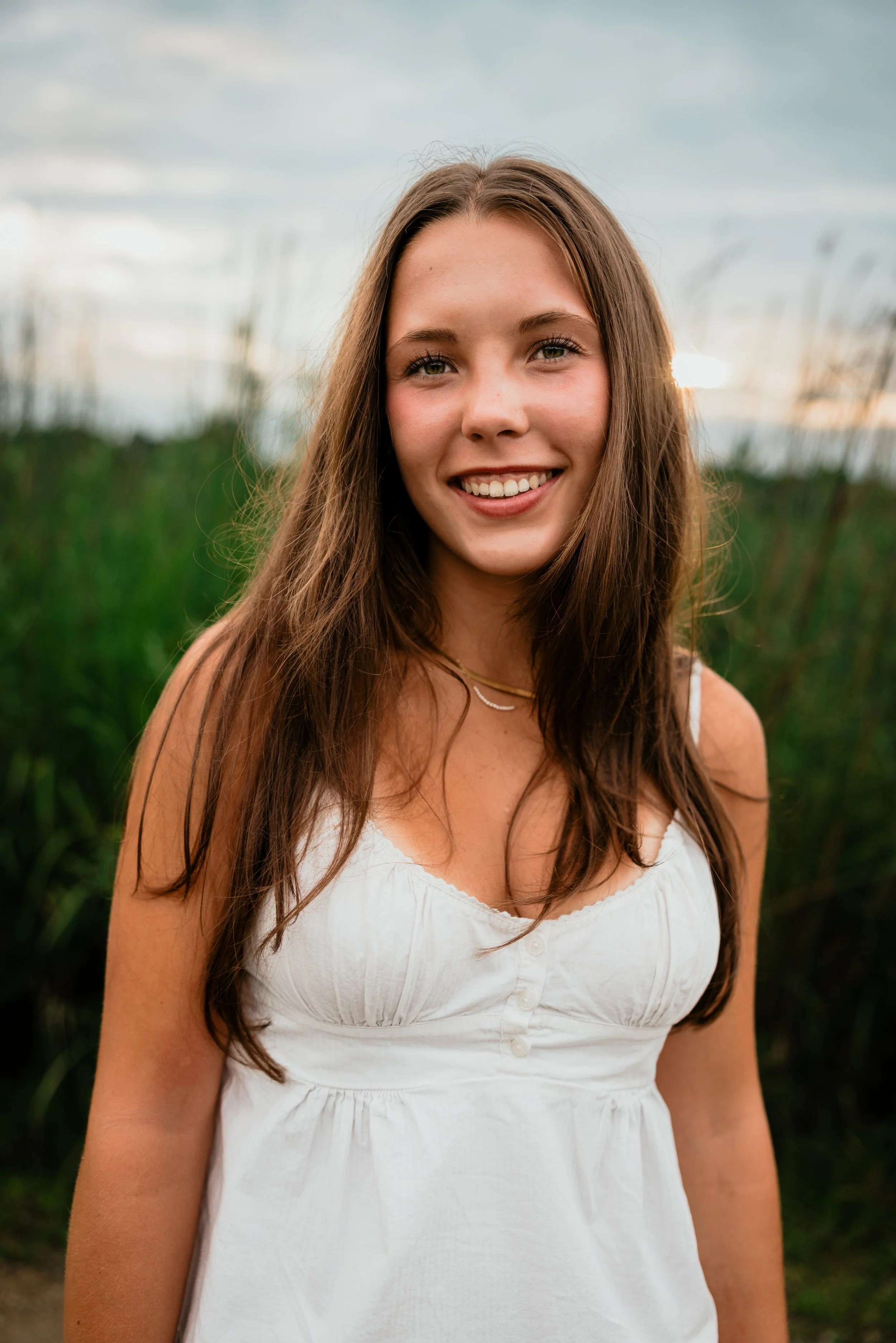 A young woman with long brown hair, smiling and wearing a white sleeveless dress, standing outdoors with green foliage and cloudy sky in the background.