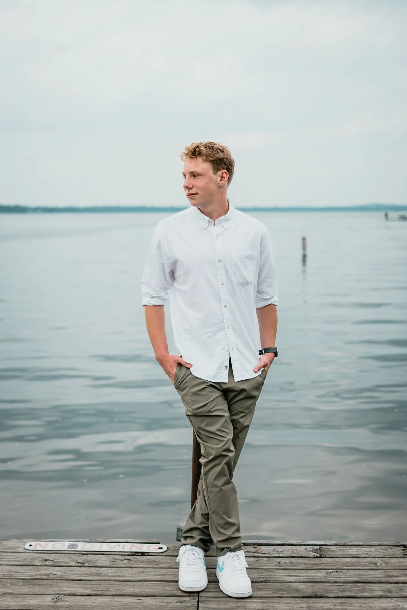 A young man with curly blond hair, wearing a white button-up shirt and khaki pants, stands on a wooden dock with his hands in his pockets, by a calm body of water with a cloudy sky in the background.