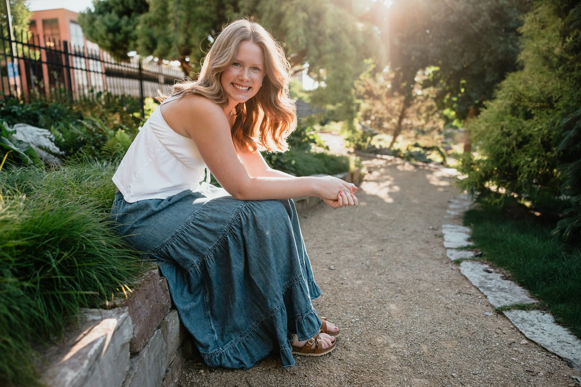 A woman with long, wavy, red hair and a white sleeveless top sitting on a stone border next to a dirt path in a sunny garden.