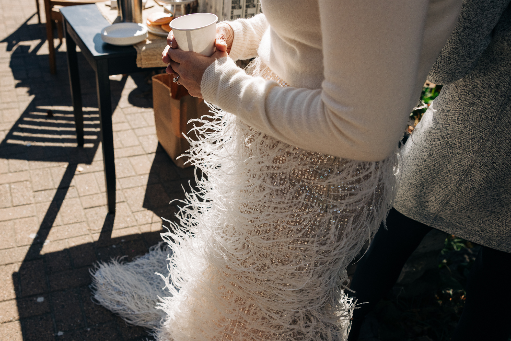 Person holding a white paper cup, standing outdoors near a black table with plates, in sunlight.