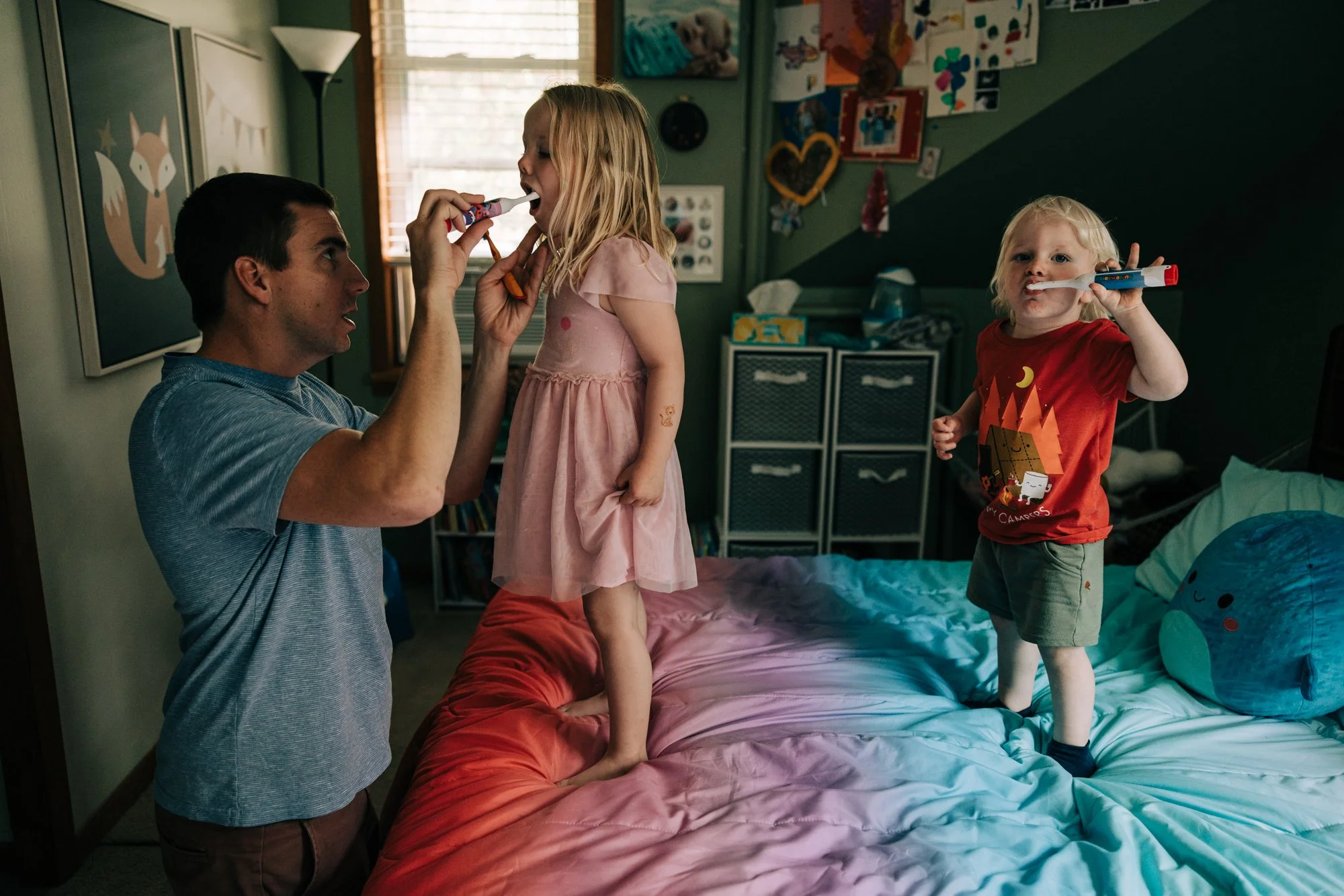 A man helps two young children brush their teeth in a bedroom, with colorful decorations on the walls and a large stuffed animal on the bed.
