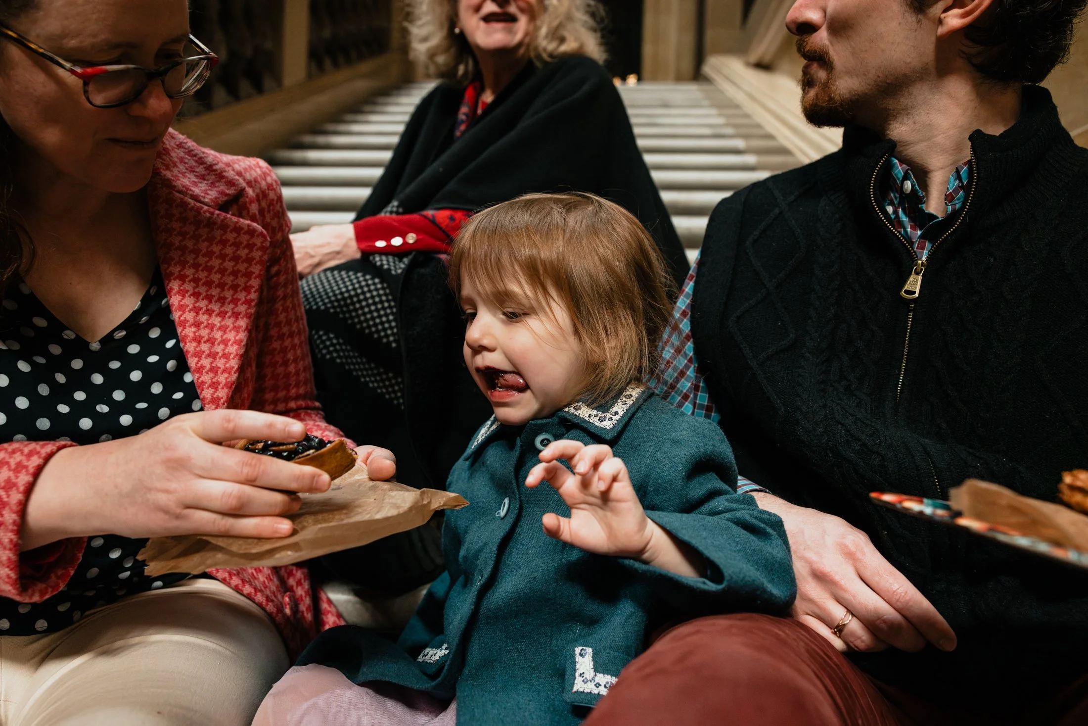 A young girl with red hair and a teal coat sits between two adults, one woman in glasses and a pink patterned jacket, and one man in a black zip-up sweater, as they share a moment together. The woman is holding a cookie with chocolate topping, while 