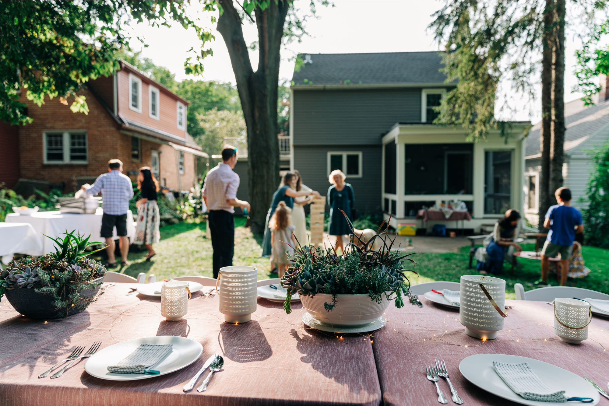 A backyard gathering with people socializing, a decorated table with candles and plants, and a house with a porch in the background.