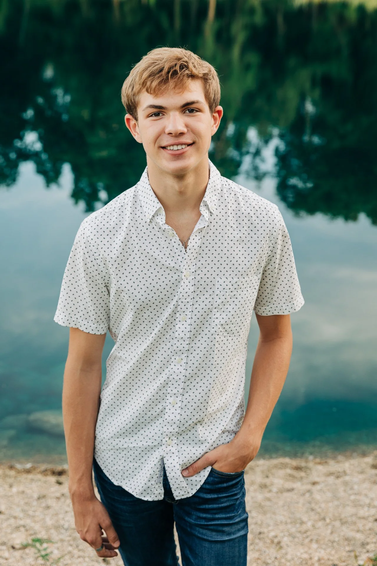 A young man with light brown hair and a white button-up shirt with small black dots, standing by a body of water with trees in the background.