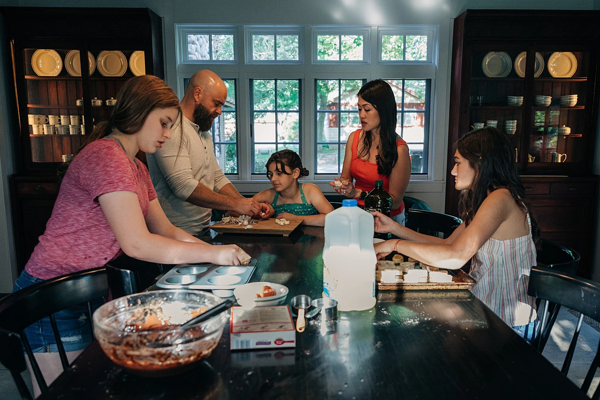 A family baking cookies in a kitchen. Five people, including a young girl, are focused on preparing cookie dough and ingredients around a table with baking supplies.
