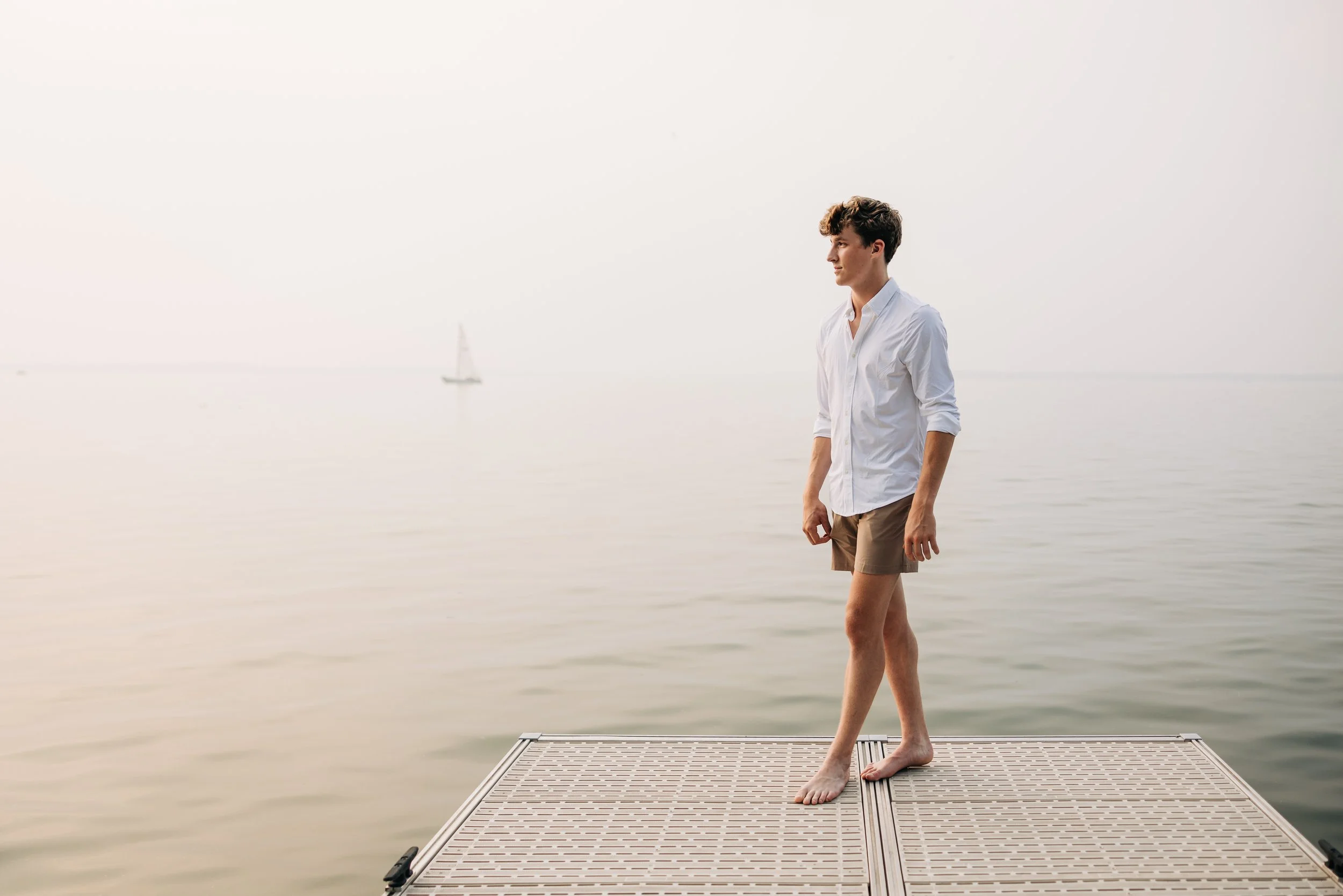 Young man standing barefoot on a dock by calm water, with a sailboat in the distance and a hazy sky.