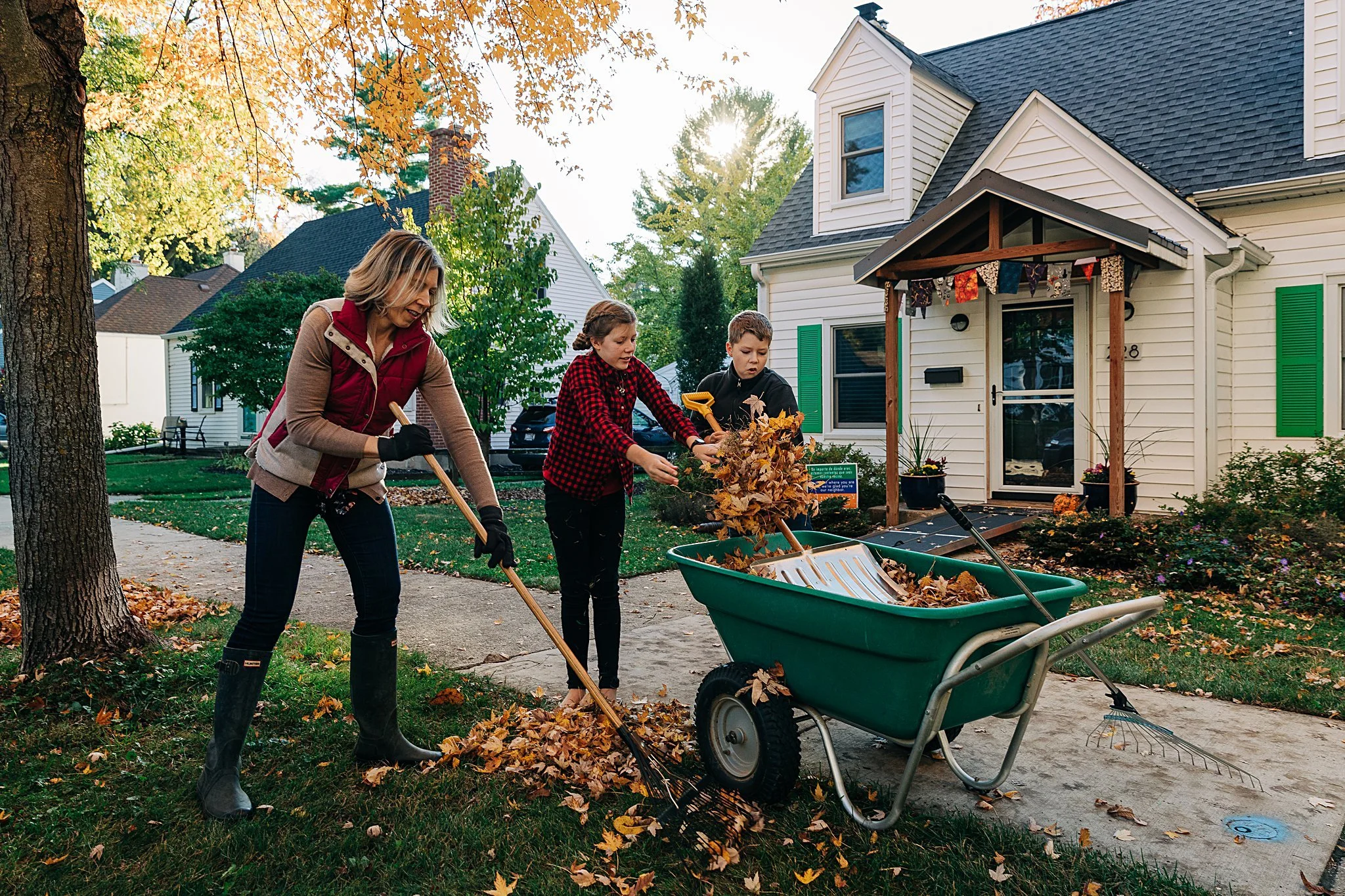 A woman and two children raking and collecting fallen leaves on a front yard lawn near a house in autumn.