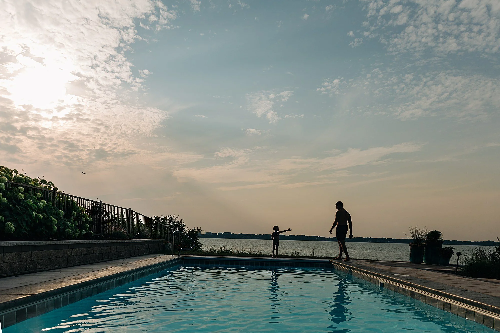 dad and daughter silhouetted against lake and sky as they enter swimming pool in foreground