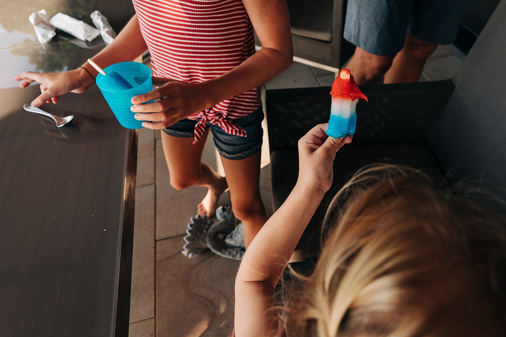 girl in red and white stripes and a blue cup and in foreground a different girl holds red white and blue popsicle