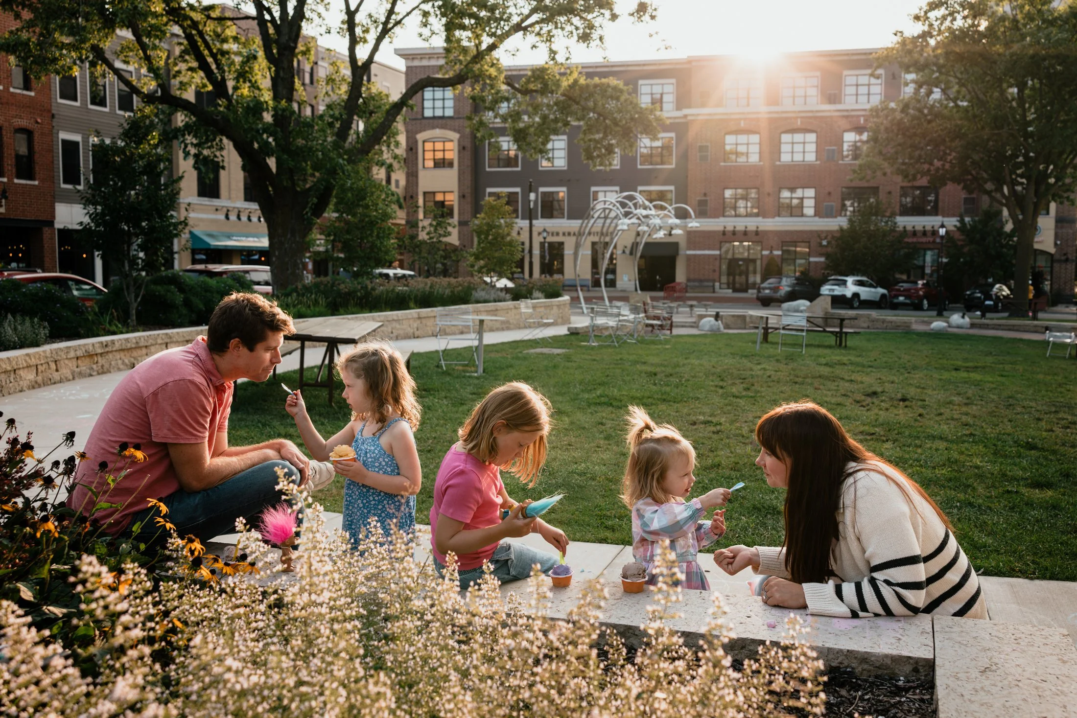A family enjoying a day at a park with children and adults having ice cream on a sunny evening, with buildings and trees in the background.